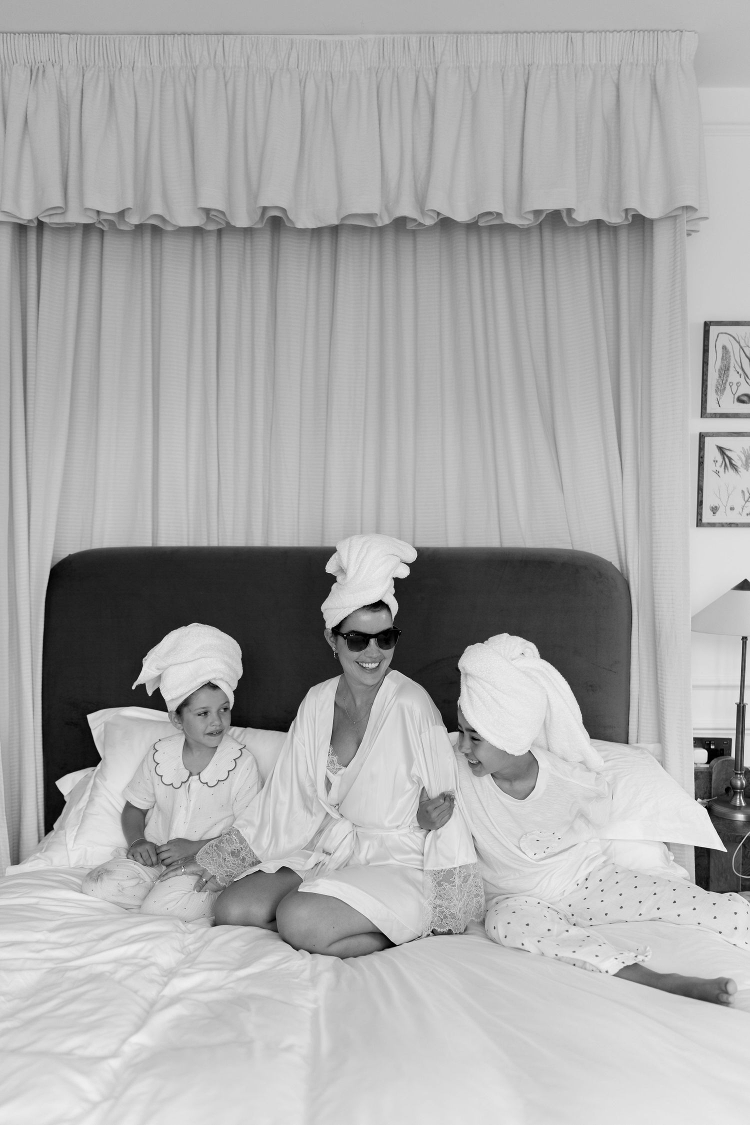 Three women in white robes and towel turbans laugh together on a bed in black and white photo.