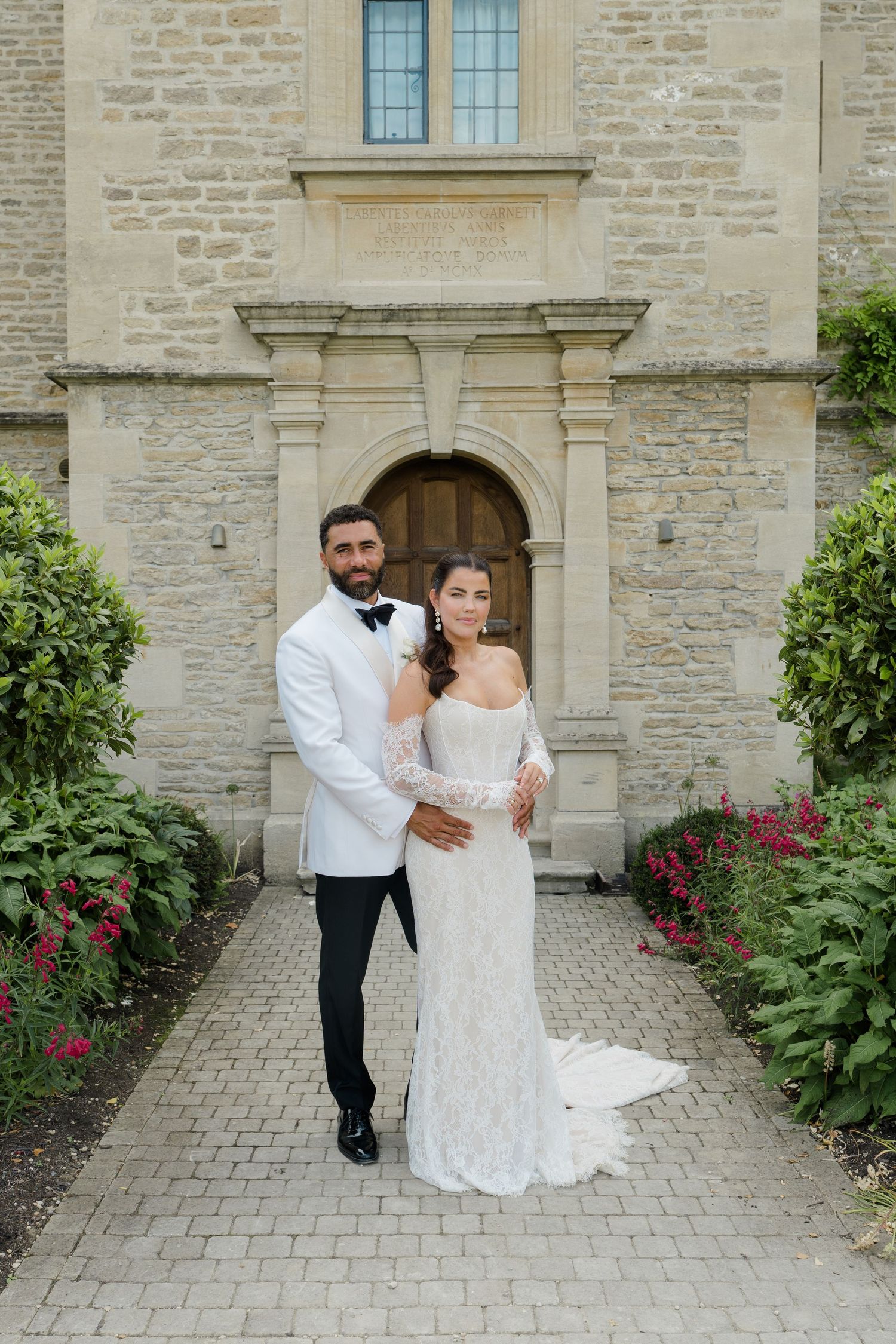 Bride and groom pose elegantly before a stone chapel, she in lace gown, he in white tuxedo jacket.