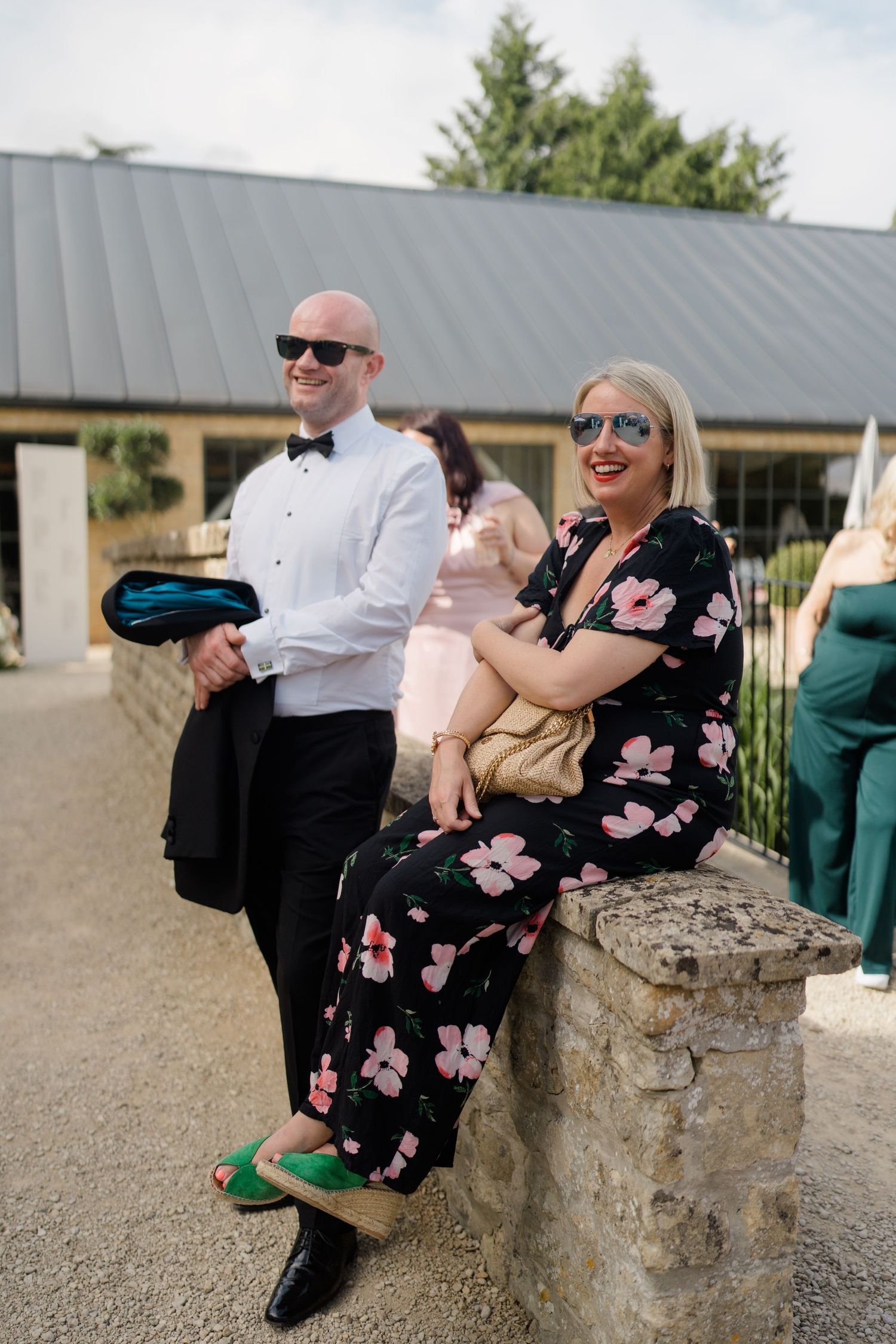Stylish wedding guests relaxing on a stone wall outside a venue, wearing sunglasses and formal attire.