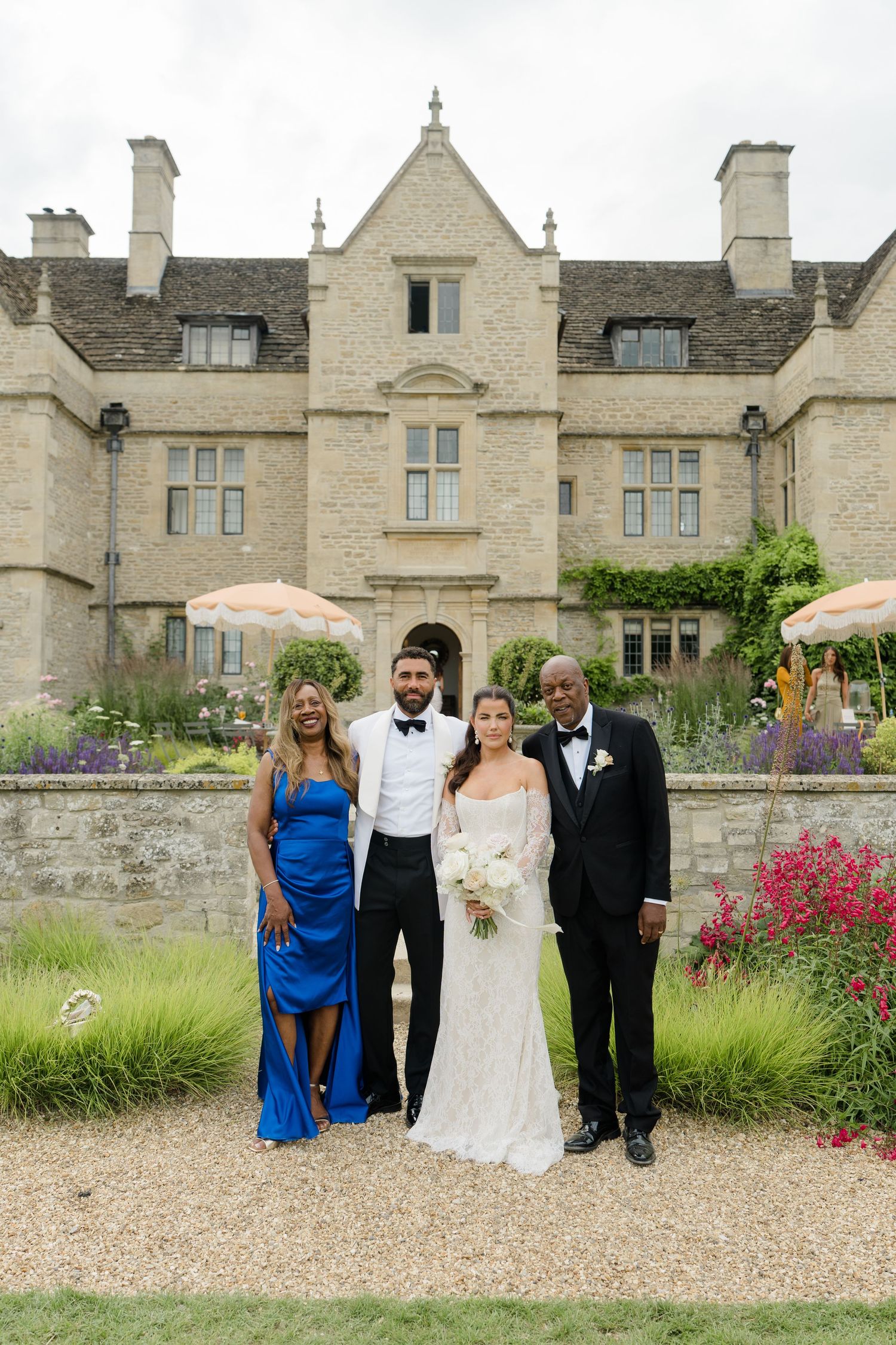 Bride in lace gown and groom in tuxedo pose with family outside a grand Cotswolds manor house.