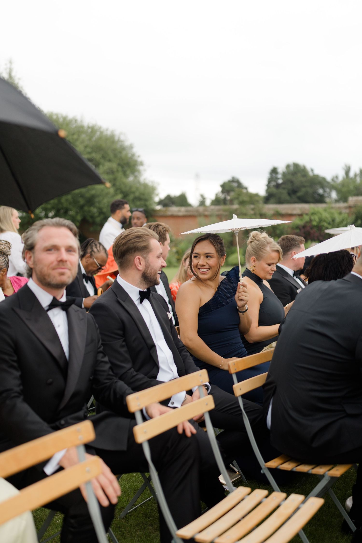 Wedding guests in formal attire seated on wooden chairs at outdoor garden ceremony with umbrellas.