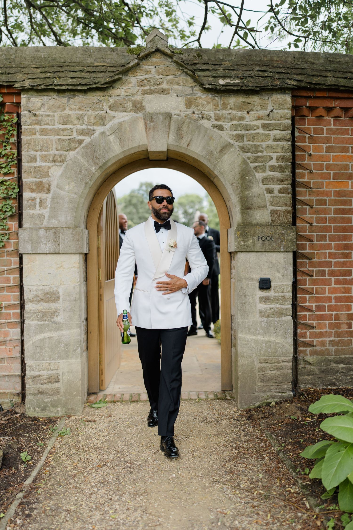 Elegant groom in a white dinner jacket walking through a stone archway at a wedding venue.