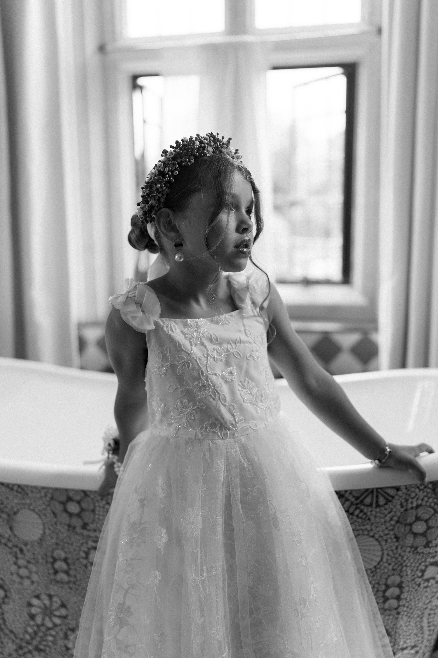 Black and white portrait of young flower girl in white dress with floral crown by bathtub.