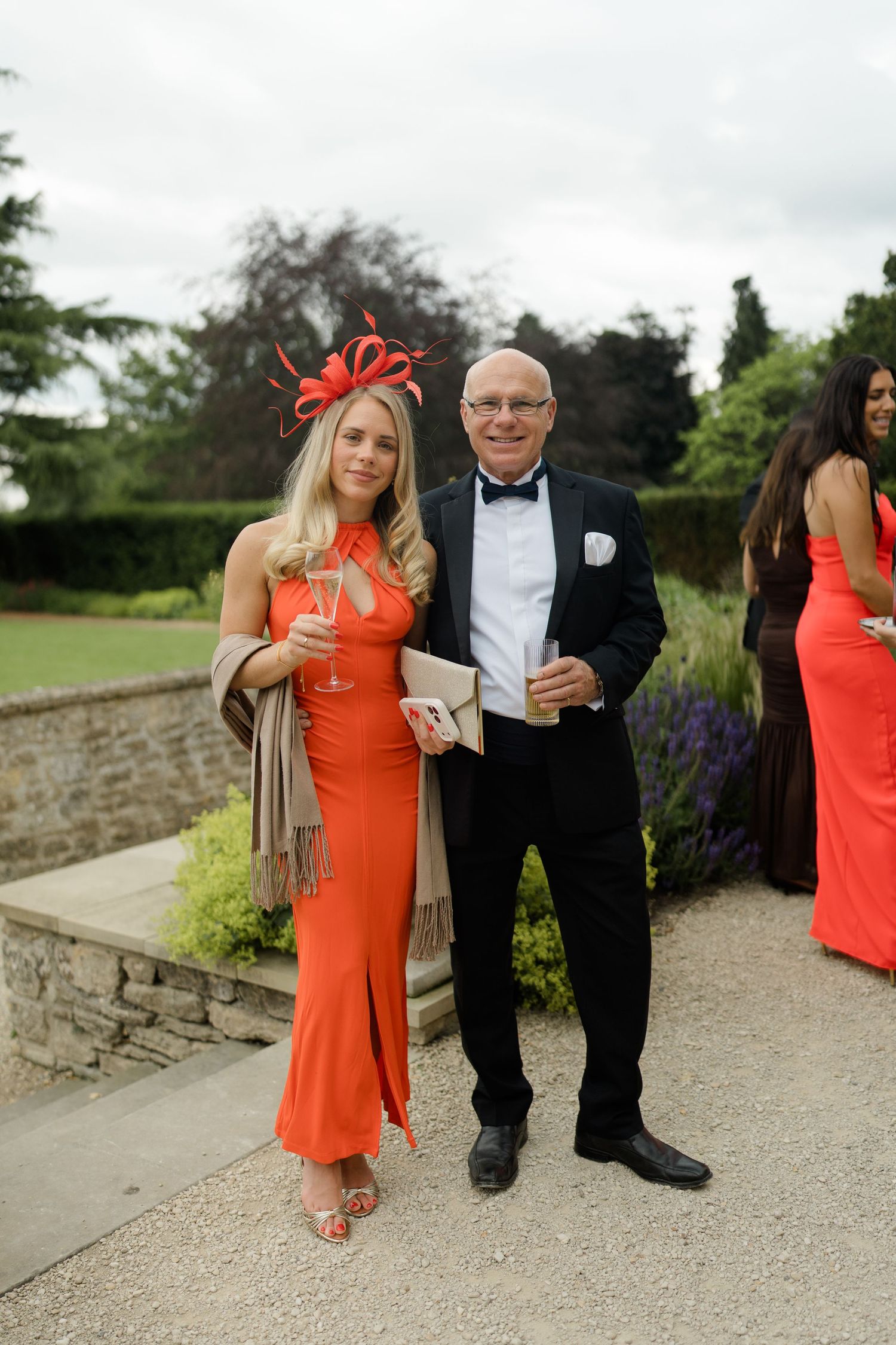 Elegant wedding guests in orange dress with fascinator and man in tuxedo posing at outdoor venue.