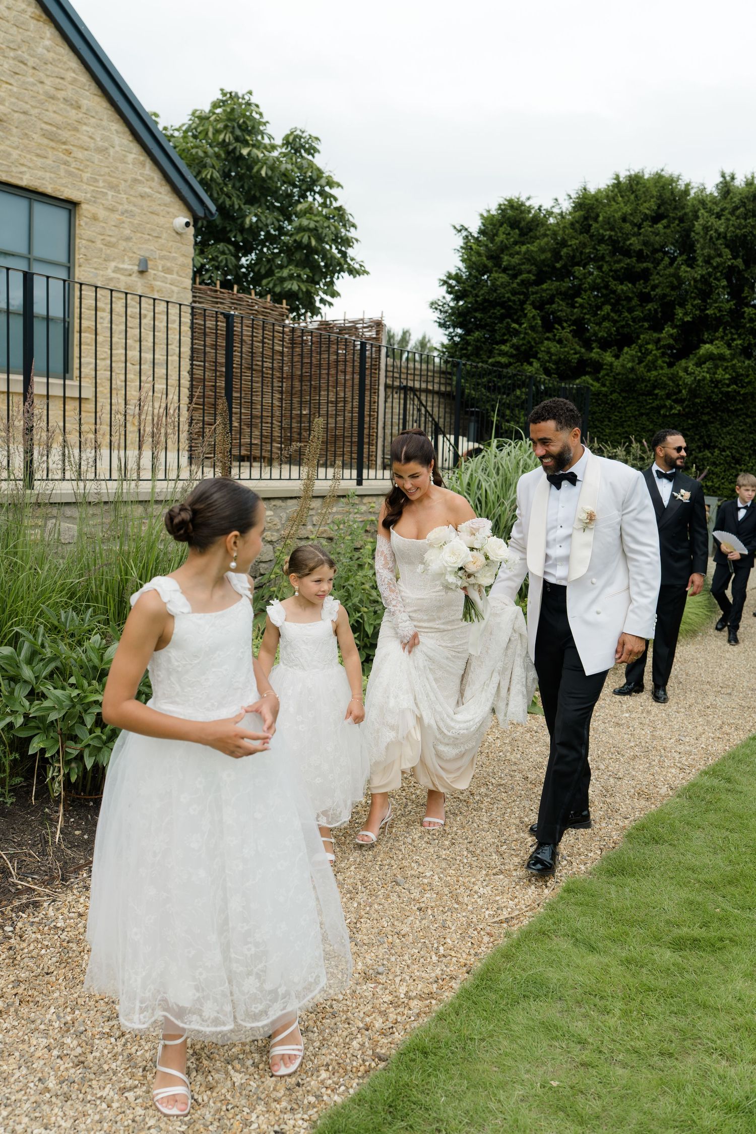Bride and groom in white jacket walk garden path with flower girls in white dresses.