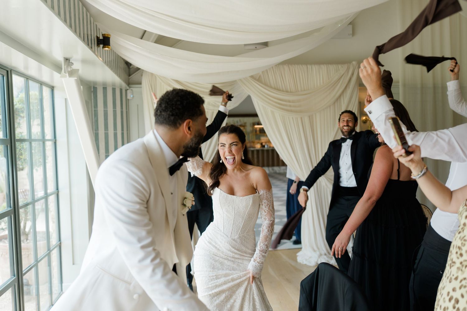 Joyful bride and groom celebrating with wedding party guests under draped fabric in bright reception venue.