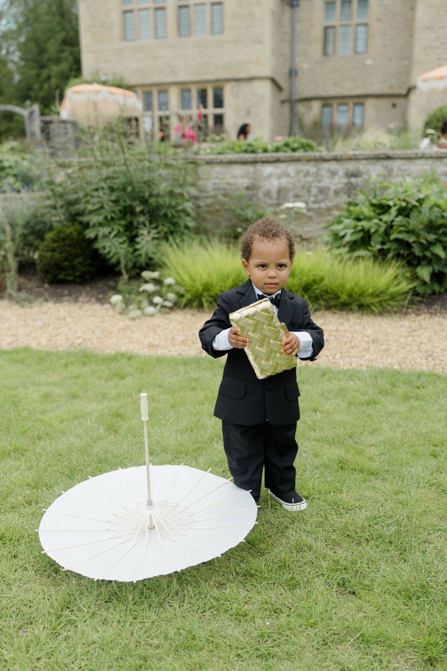 A toddler in a black tuxedo holds a decorative box beside a white parasol on a wedding venue lawn.