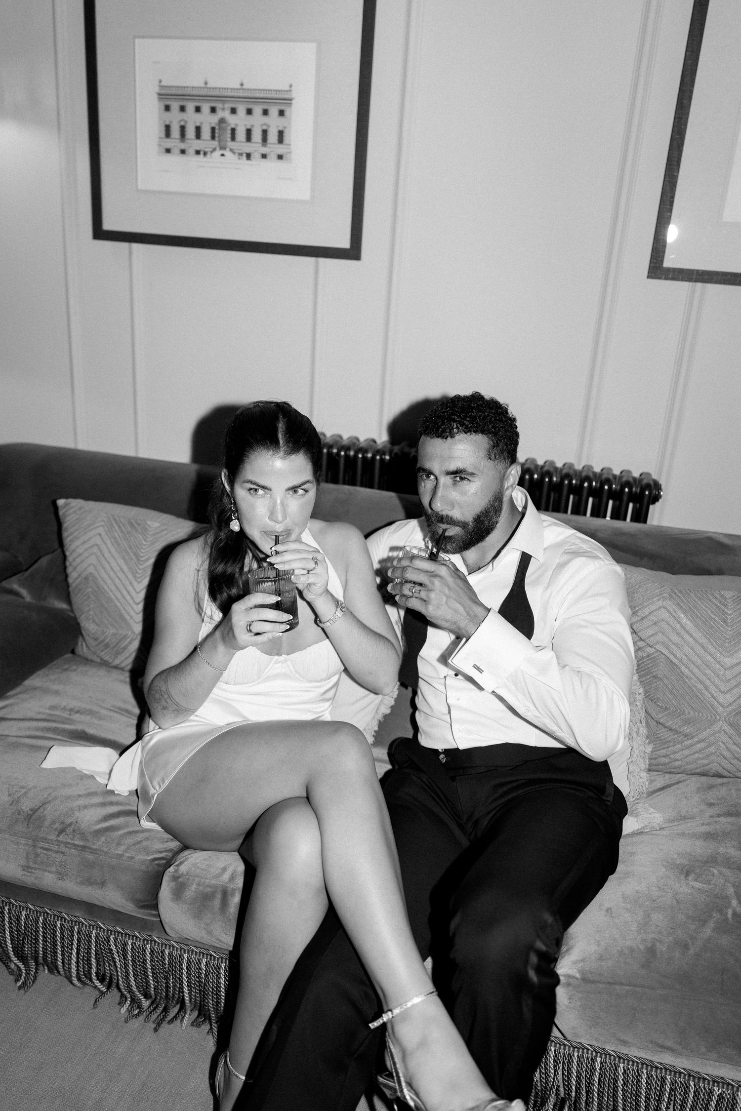 Black and white photo of elegant couple in formal wear sipping drinks together on velvet sofa.