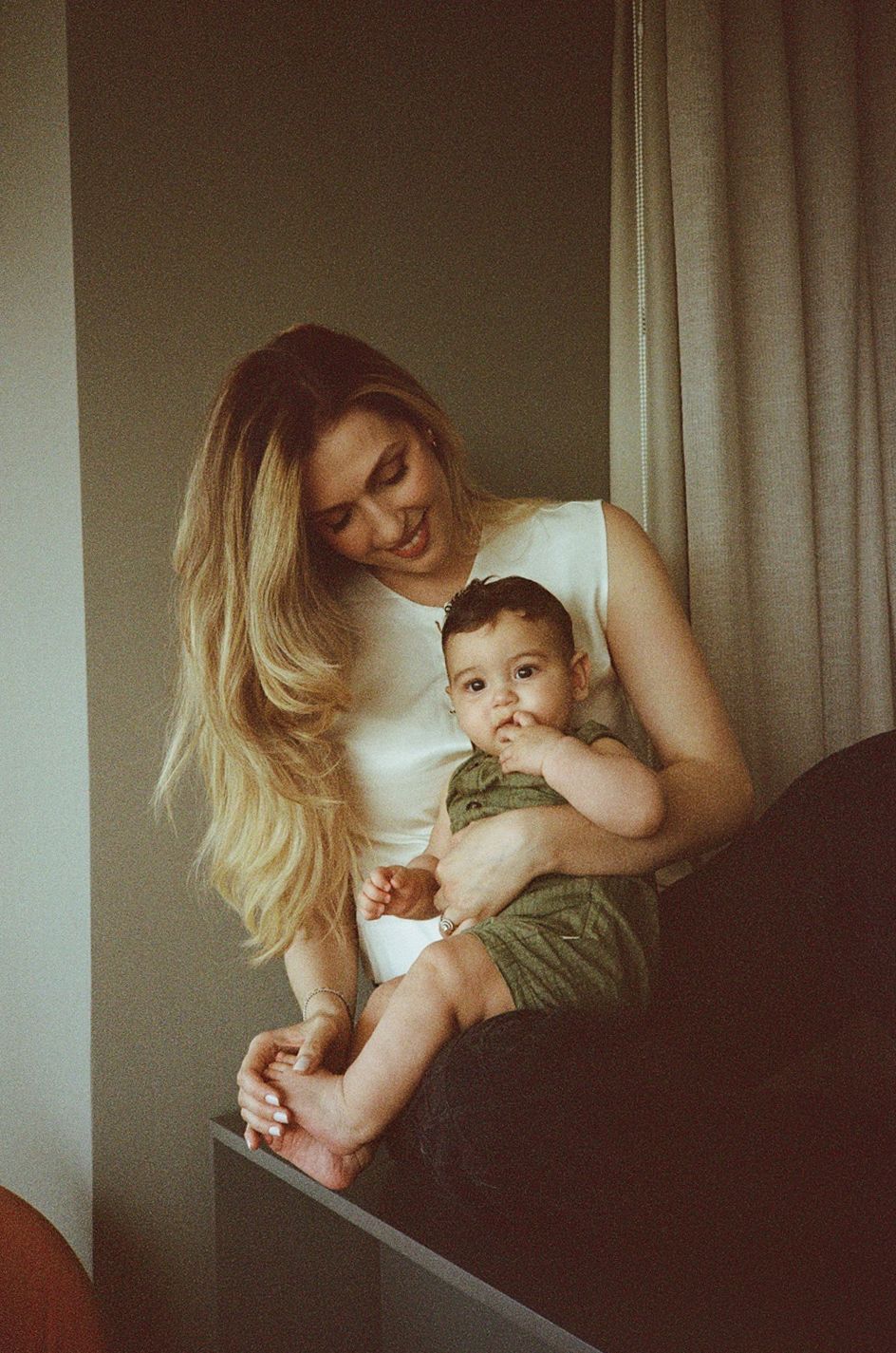 A smiling blonde woman holds a cute baby in green shorts near a window with soft curtains.