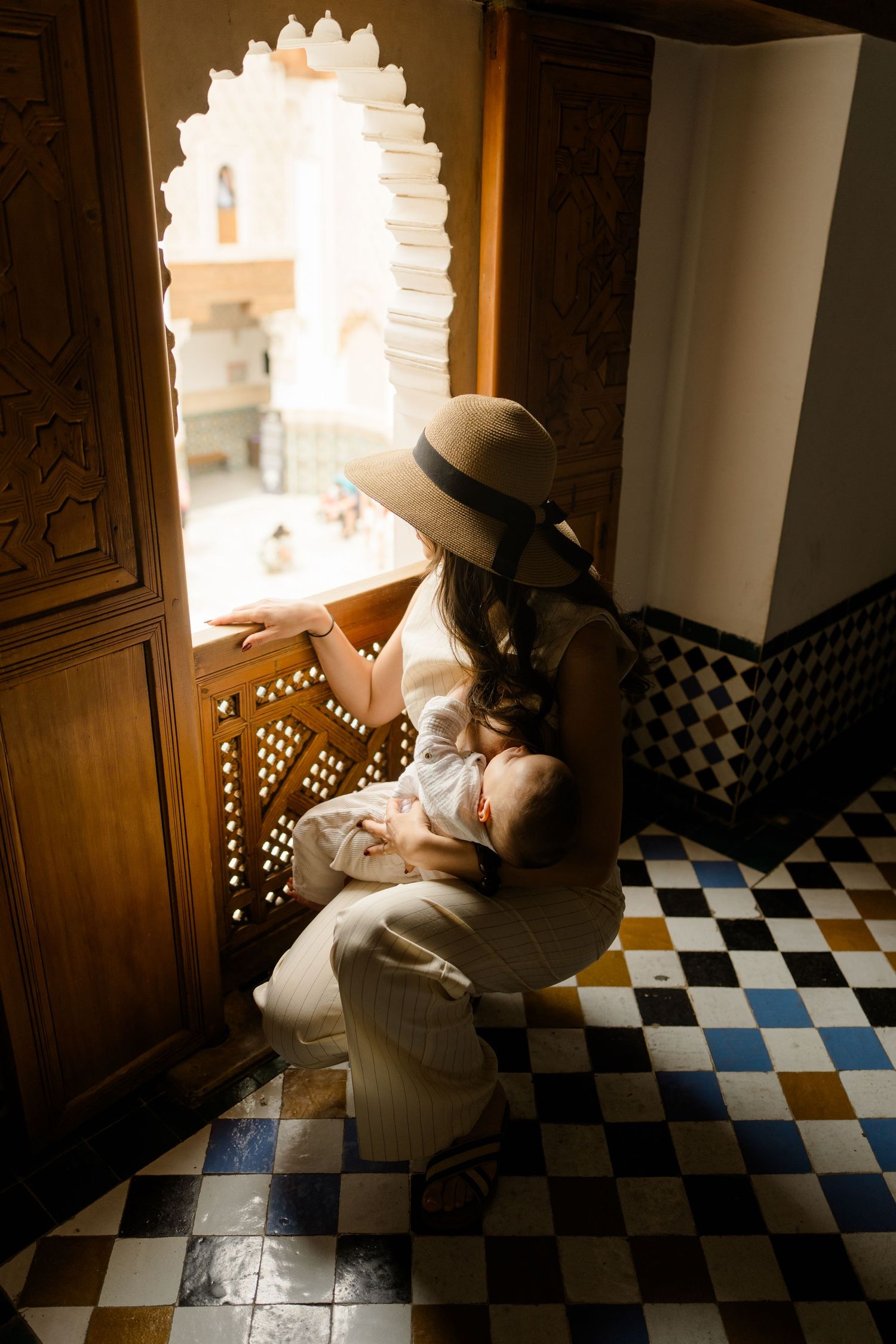 Mother in sun hat breastfeeding baby by ornate Moroccan window with geometric tilework floor.