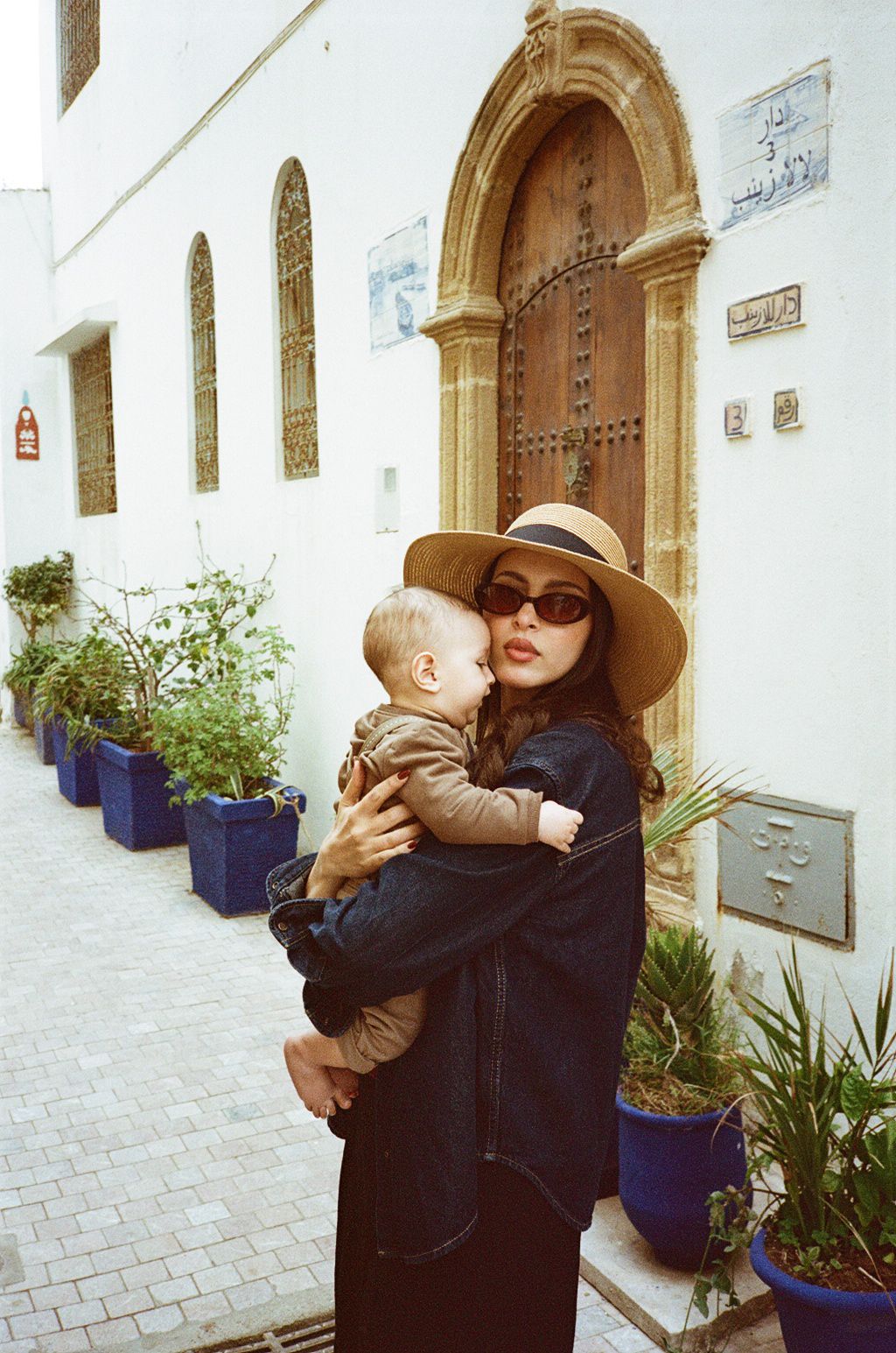 Woman in wide-brim hat and sunglasses holding a baby outside a Moroccan building with blue planters.