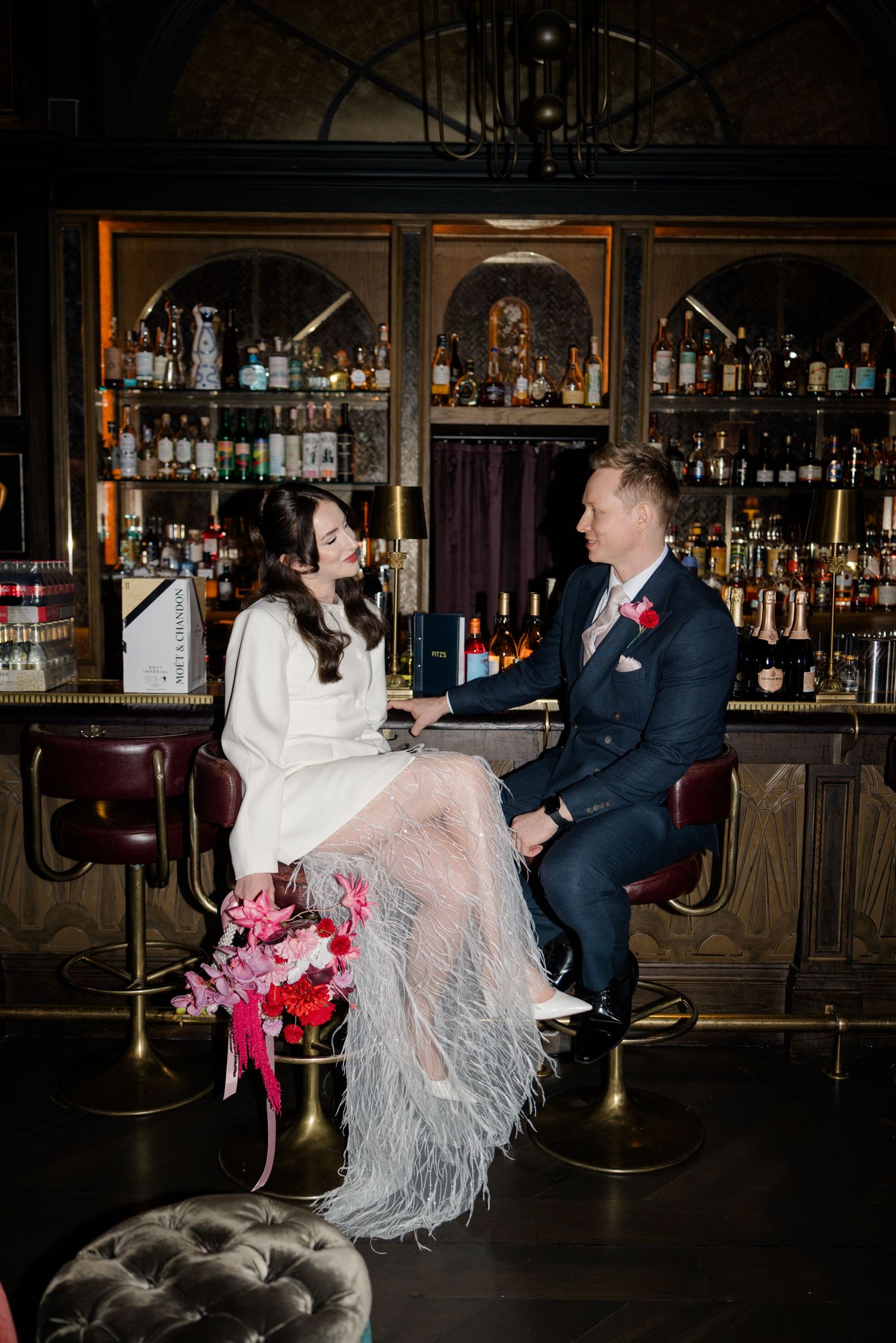 Bride and groom sitting at a dark bar together, smiling and holding hands.