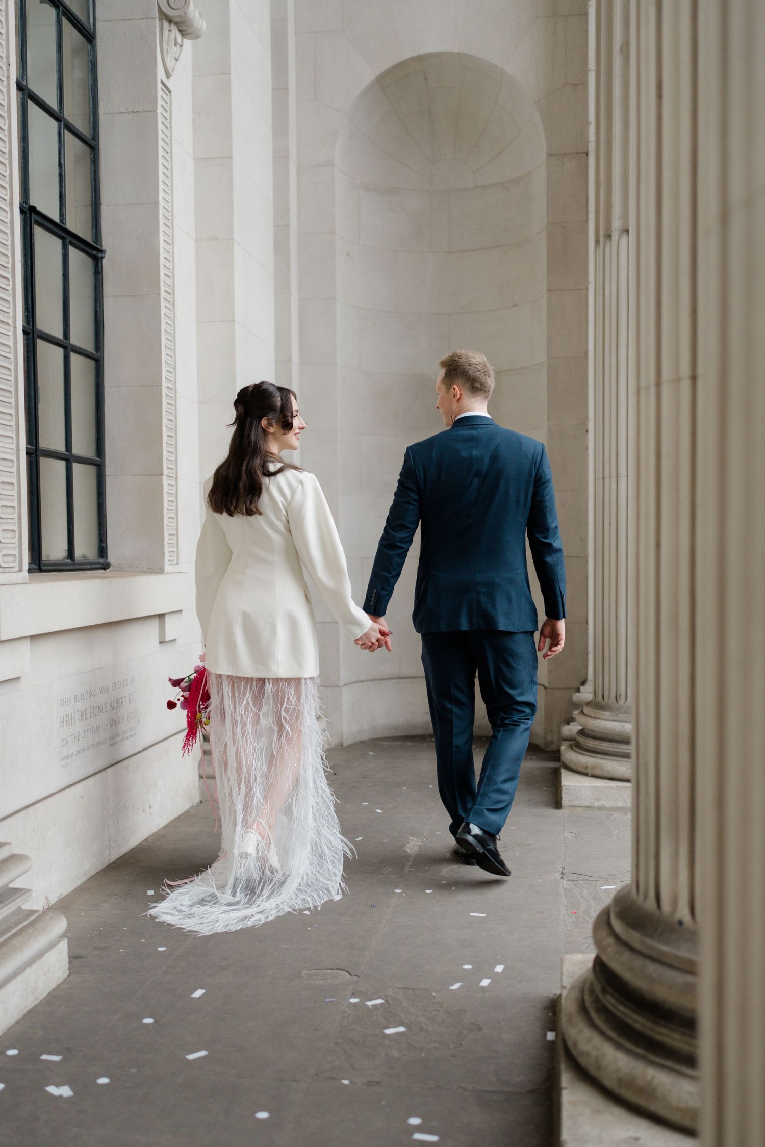 Bride in white feather-trimmed dress and groom in navy suit walk hand in hand past stone columns.