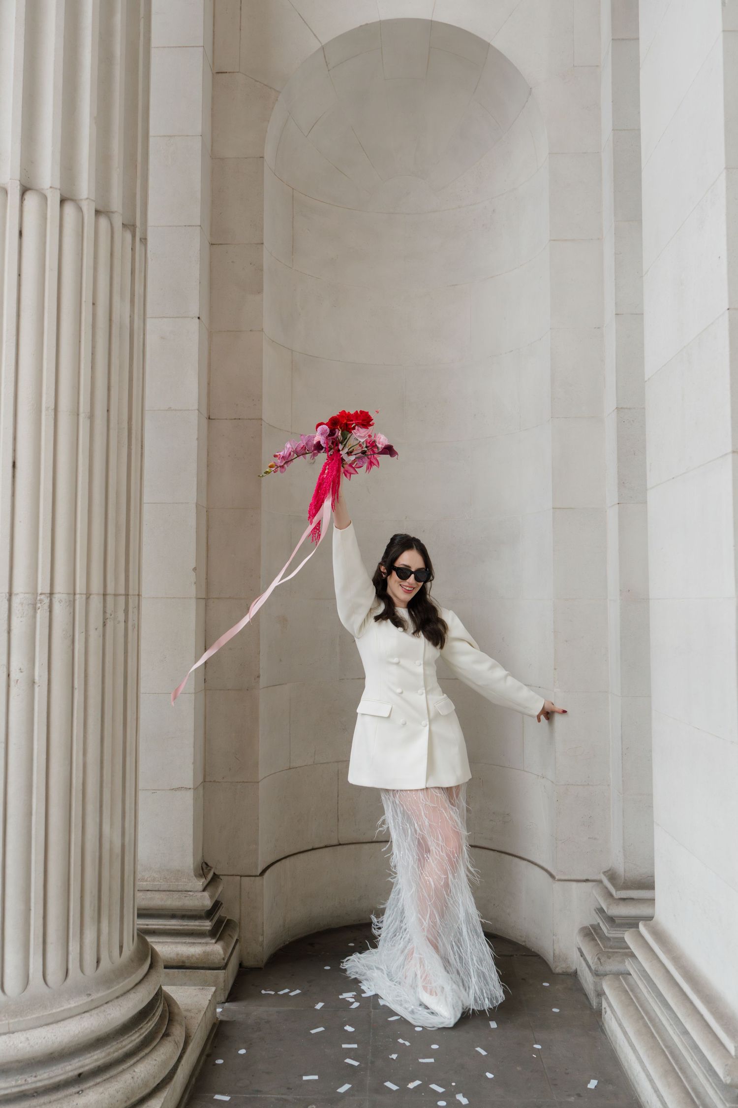 Bride in white mini dress with feather skirt raises pink floral bouquet between classical stone columns.