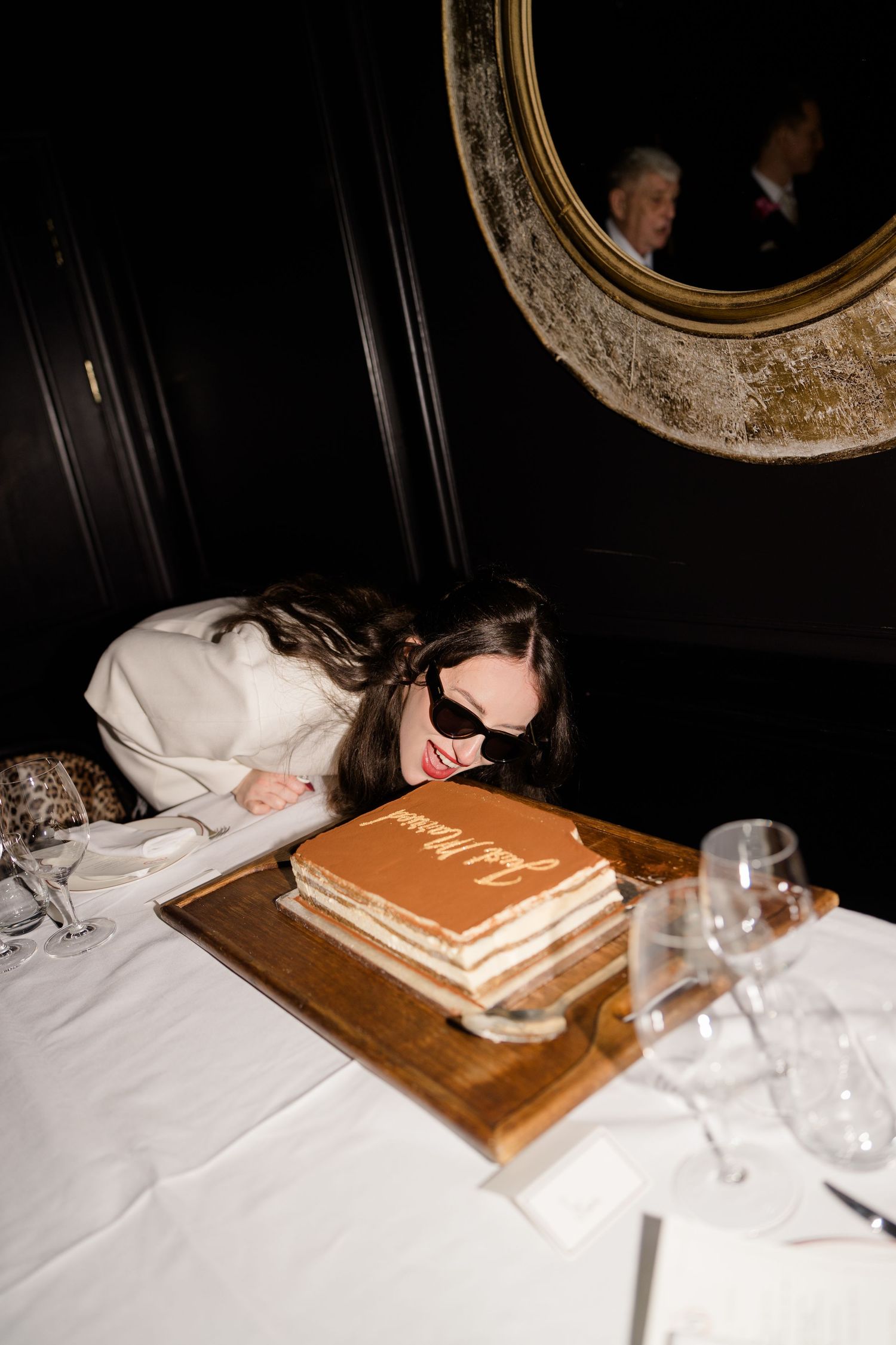 Bride leaning over a decorated wedding cake with sunglasses at a dark elegant restaurant.