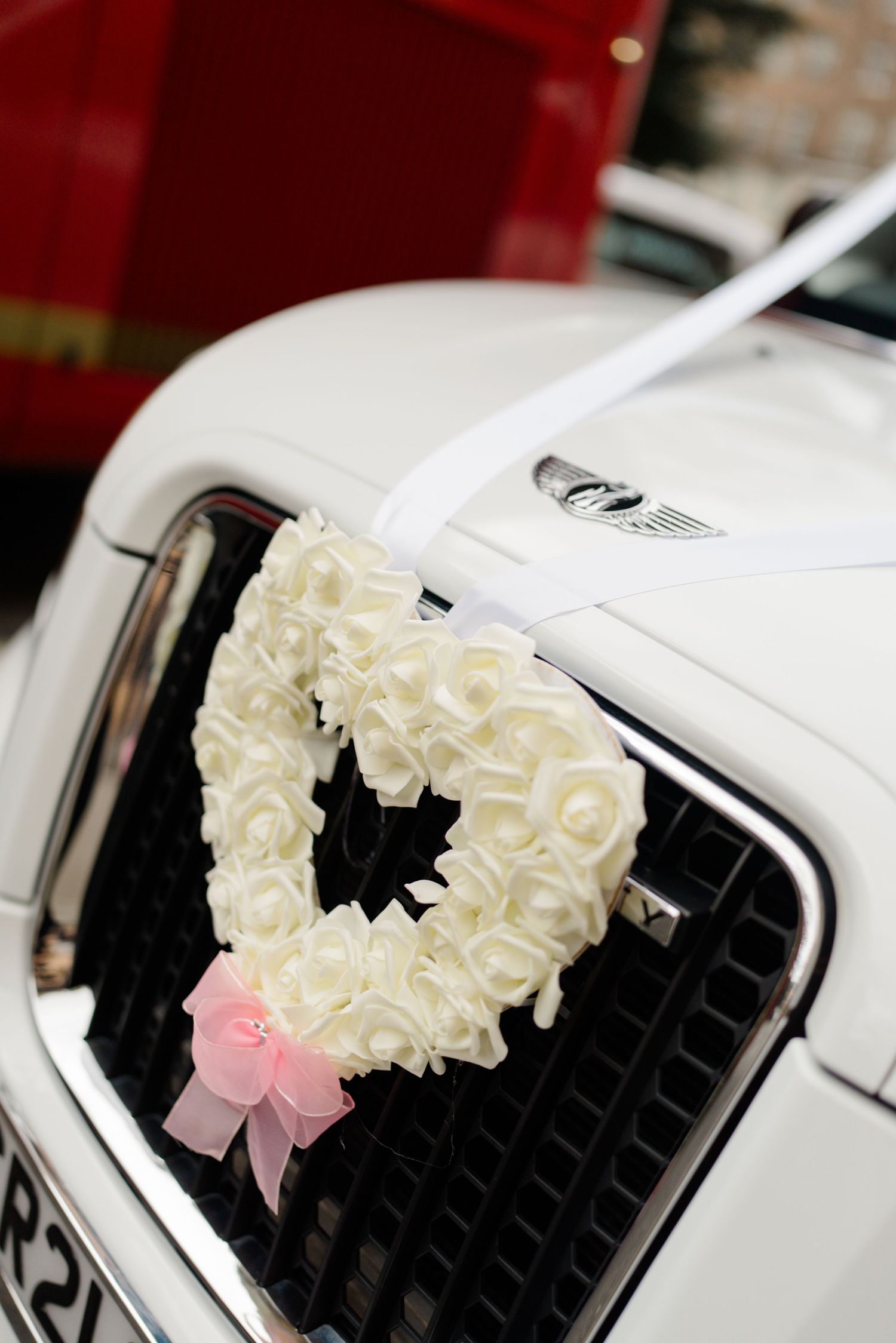 White heart-shaped floral wreath with cream roses and pink bow decorates the grille of a wedding car.