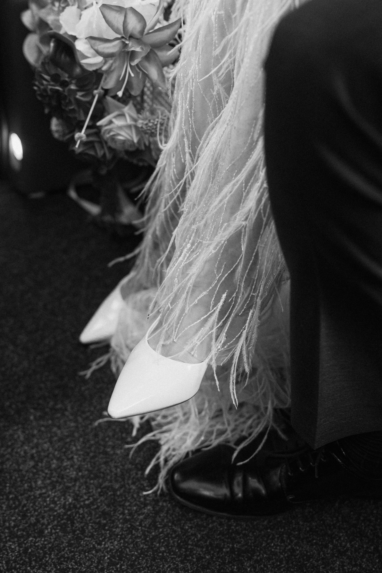 Black and white close-up of bride's white pointed heels with feathered dress hem and bouquet.