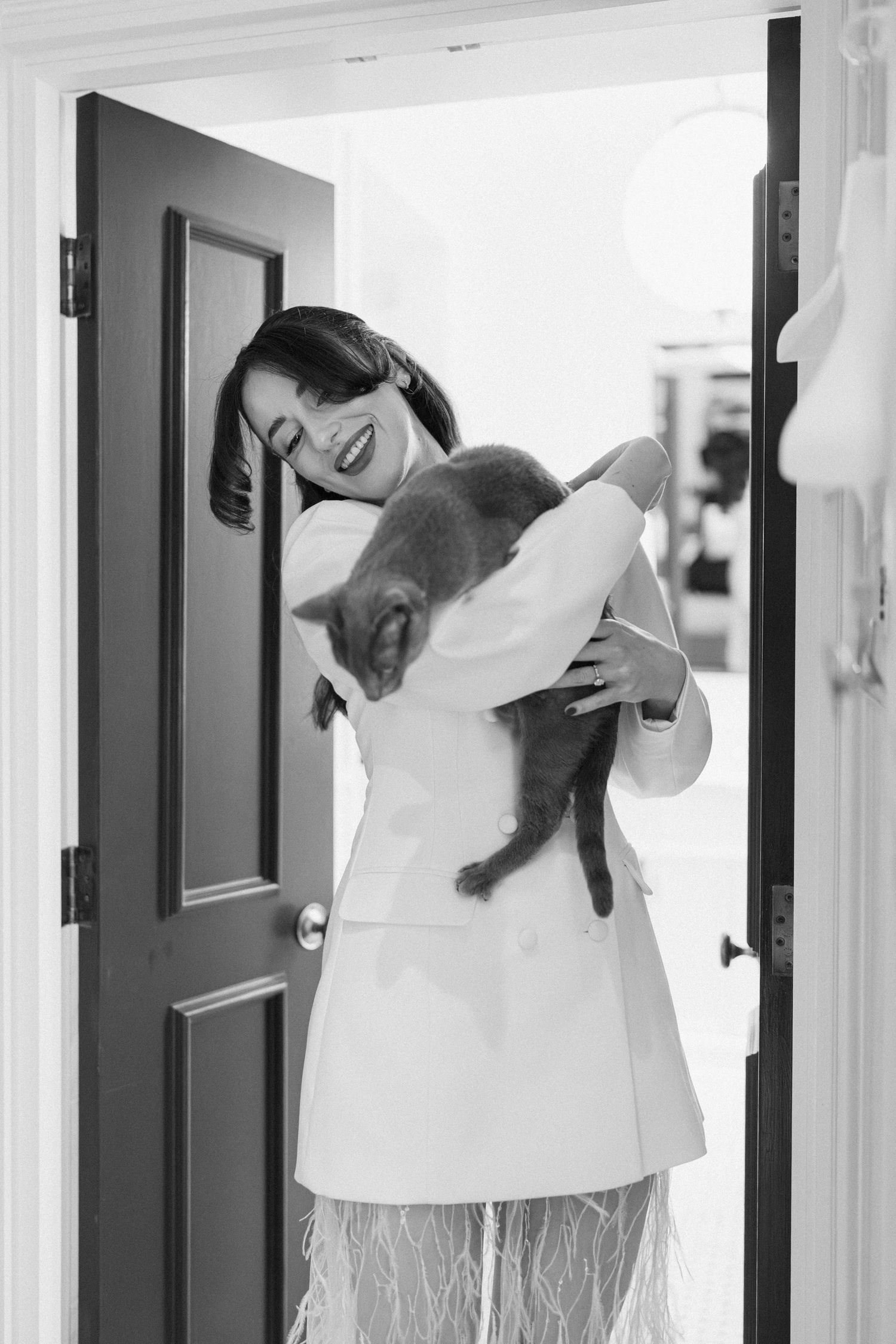 A joyful bride in a white blazer and feathered skirt hugs a grey cat in a doorway in black and white.