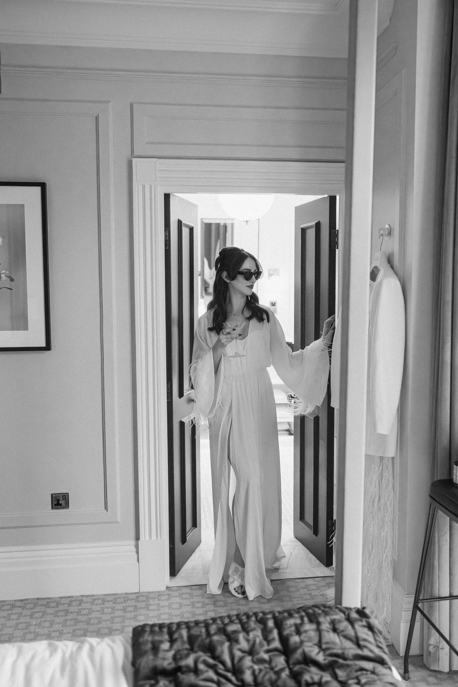 Bride in white flowing gown and sunglasses stands in hotel room doorway in black and white photo.