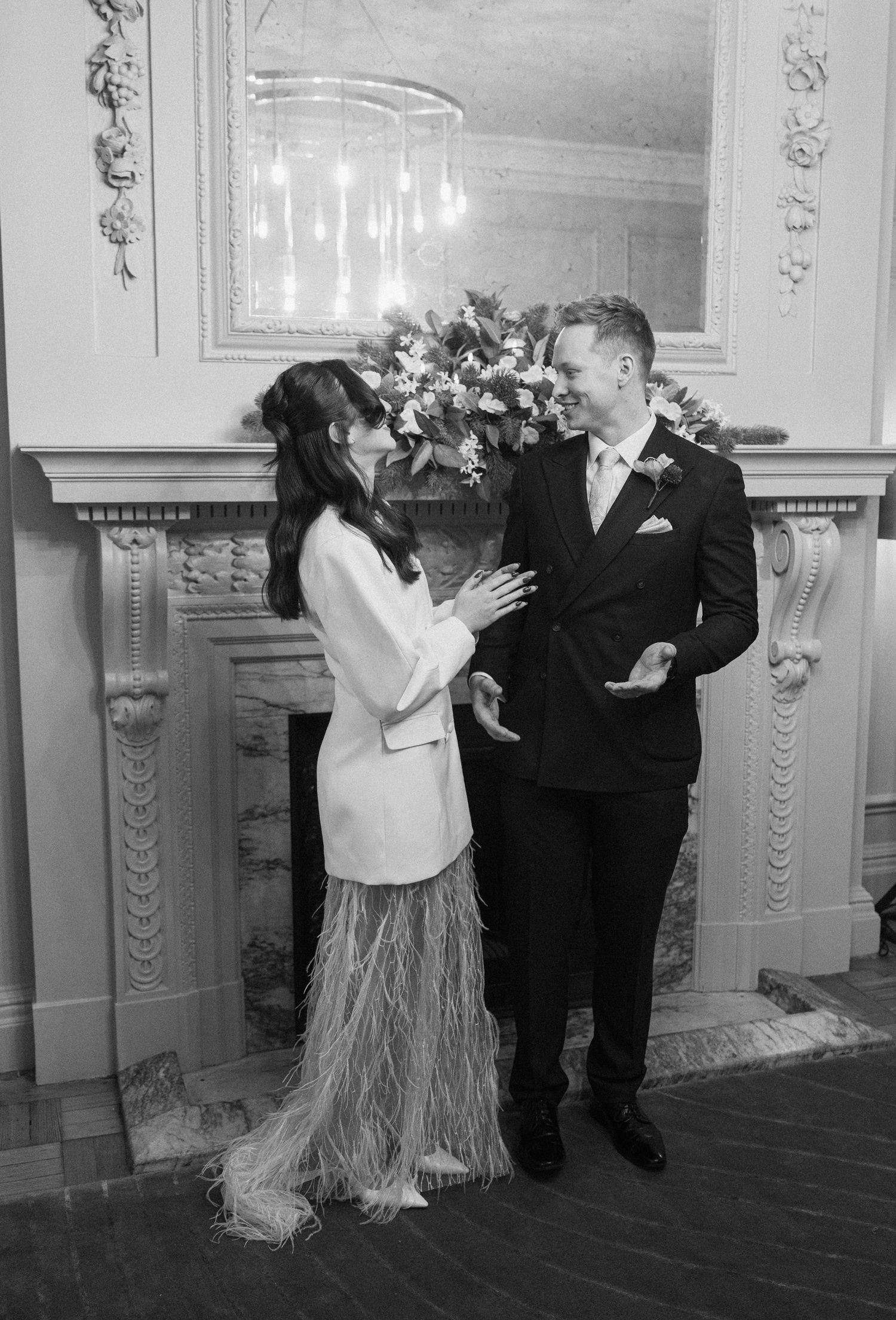 Black and white photo of bride and groom sharing a joyful moment by an elegant fireplace with floral arrangement.