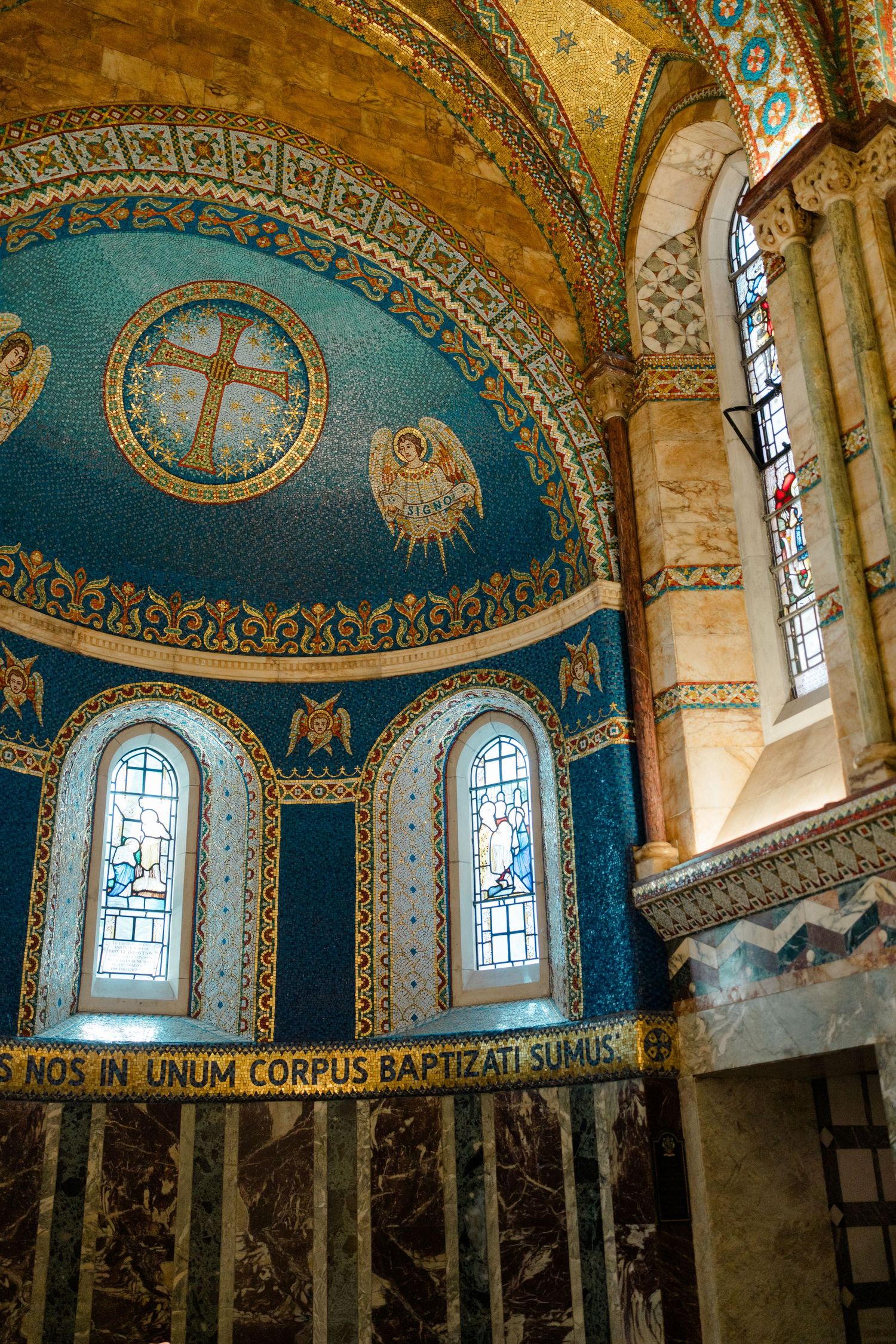 Byzantine-style church interior featuring gold mosaic apse with cross, angels, and Latin inscription above marble altar.
