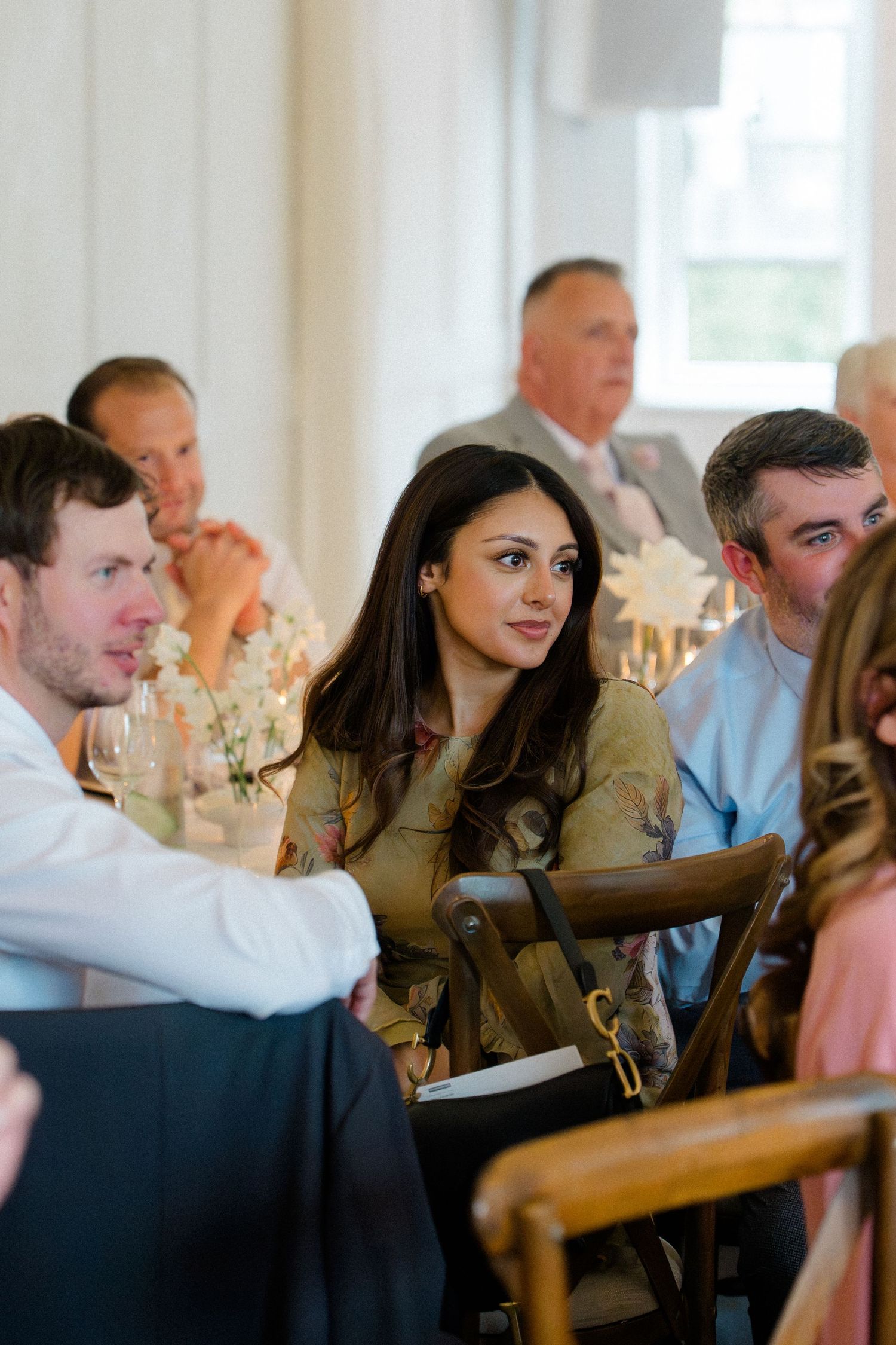 Wedding guests seated at reception tables listening attentively, with a dark-haired woman in a floral dress in focus.