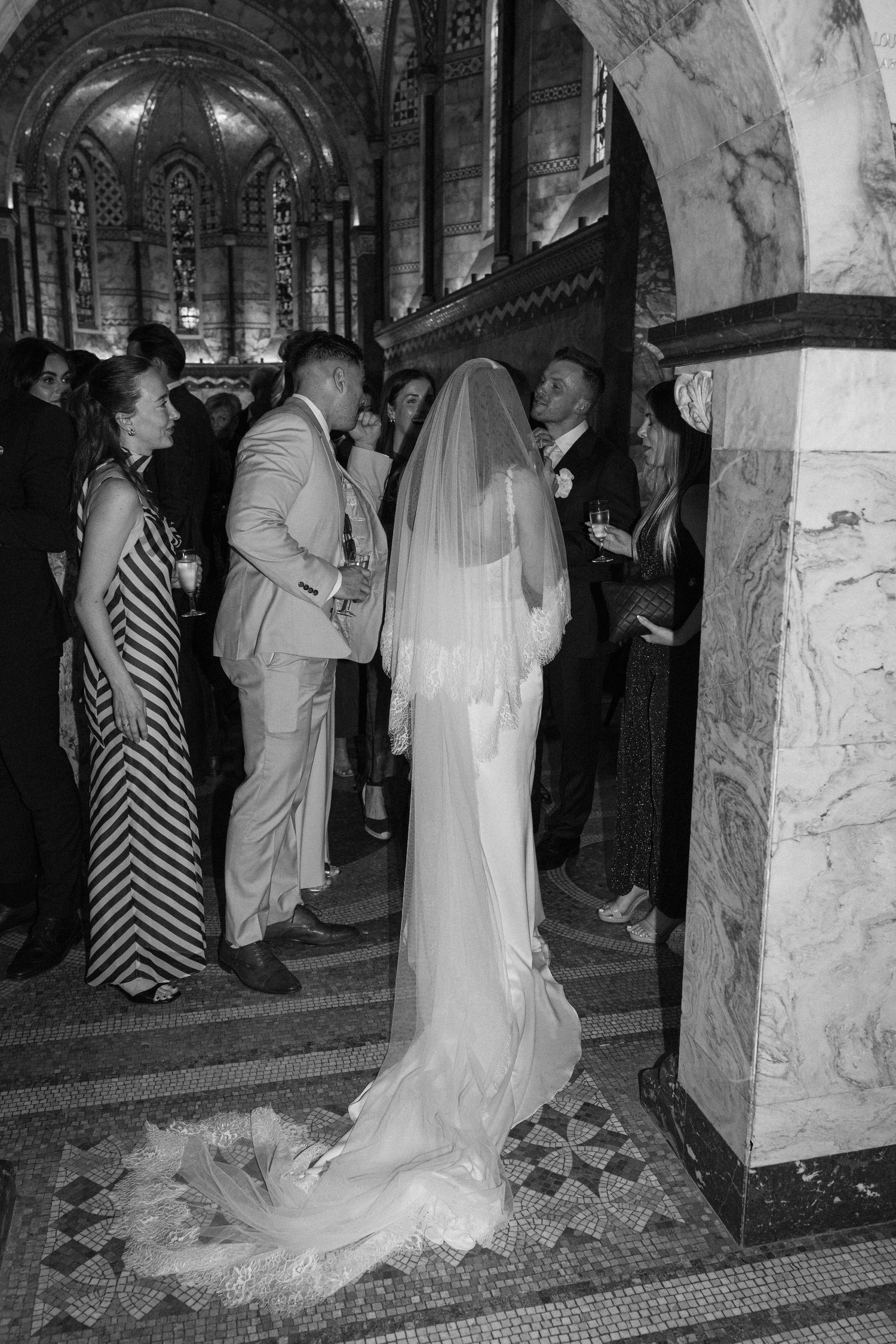 Bride in flowing white gown and cathedral veil greeting guests in ornate marble church interior.