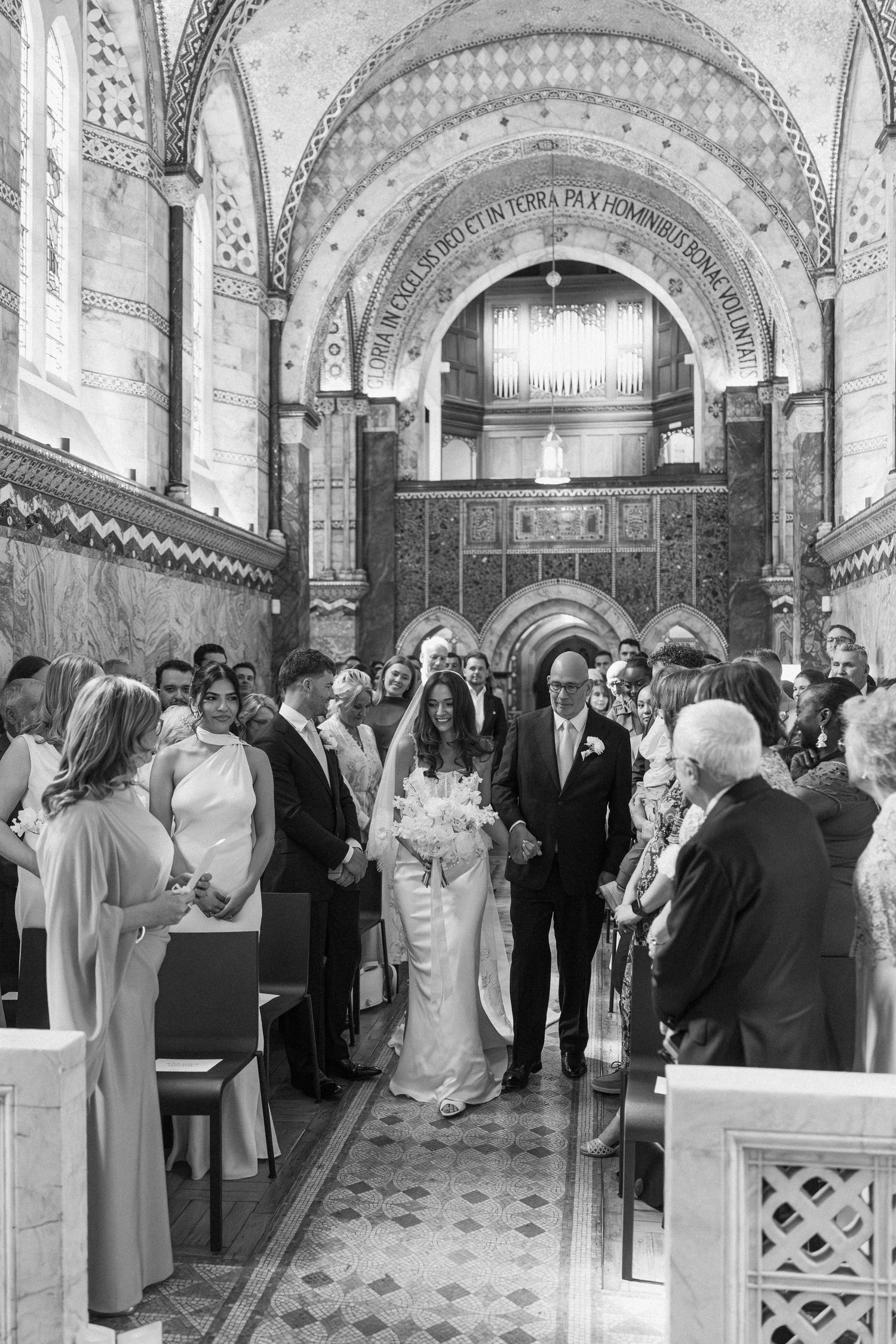 Bride walking down ornate church aisle with escort in black and white wedding ceremony photo.