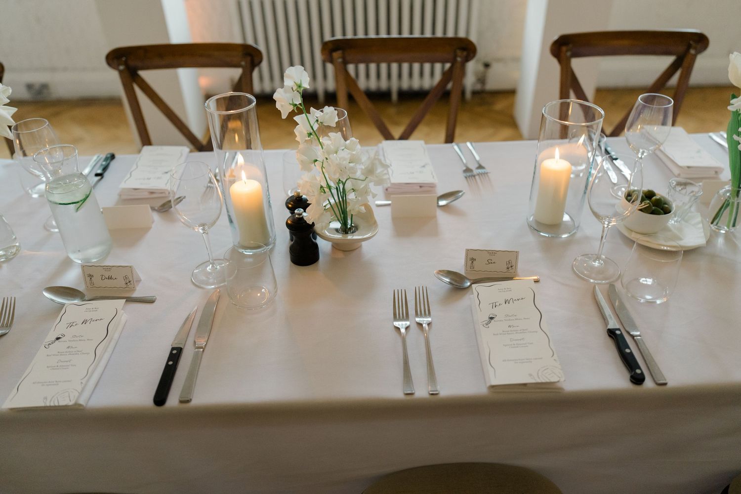 Elegant wedding reception table set with white linens, candles, glassware, menus, and white floral centerpiece.