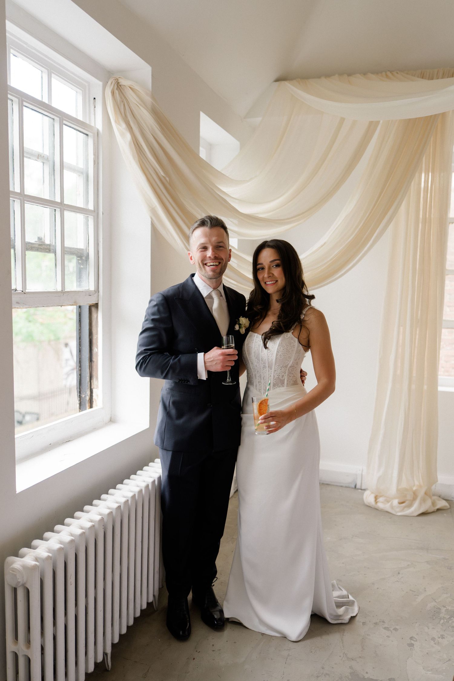 Smiling bride and groom holding champagne glasses stand before cream draped fabric at wedding reception.