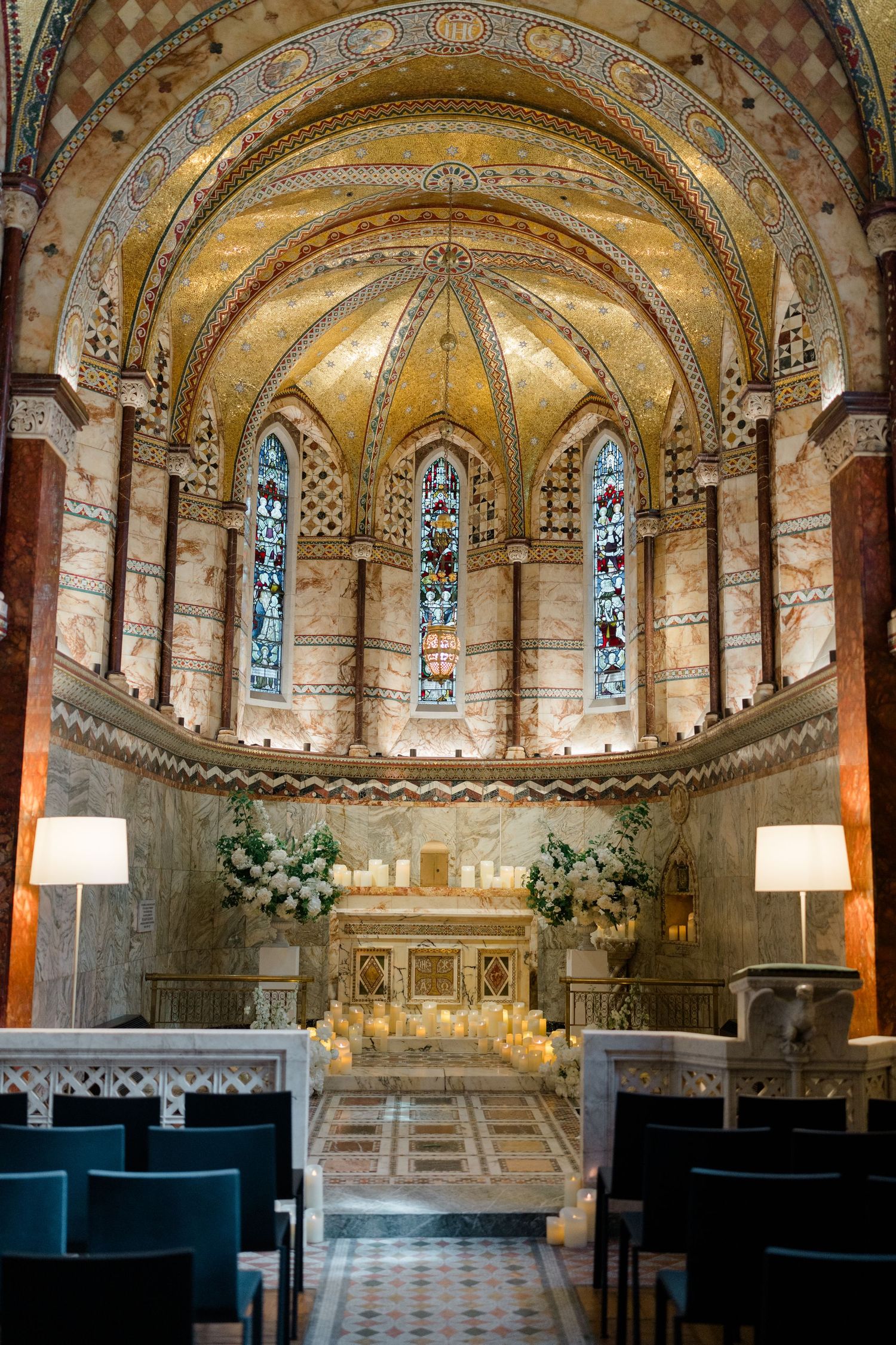 Ornate Byzantine chapel interior with golden mosaic apse, stained glass windows, and white floral arrangements for a wedding ceremony.