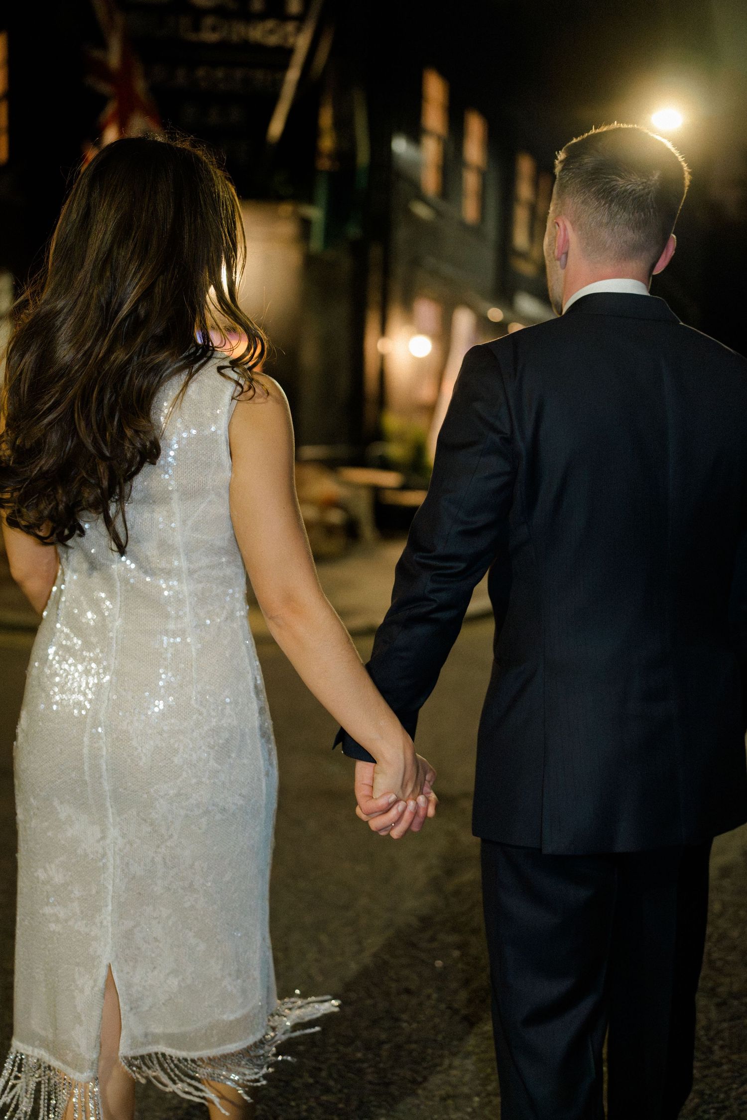 Bride in sequined white dress and groom in dark suit holding hands walking down a street at night.