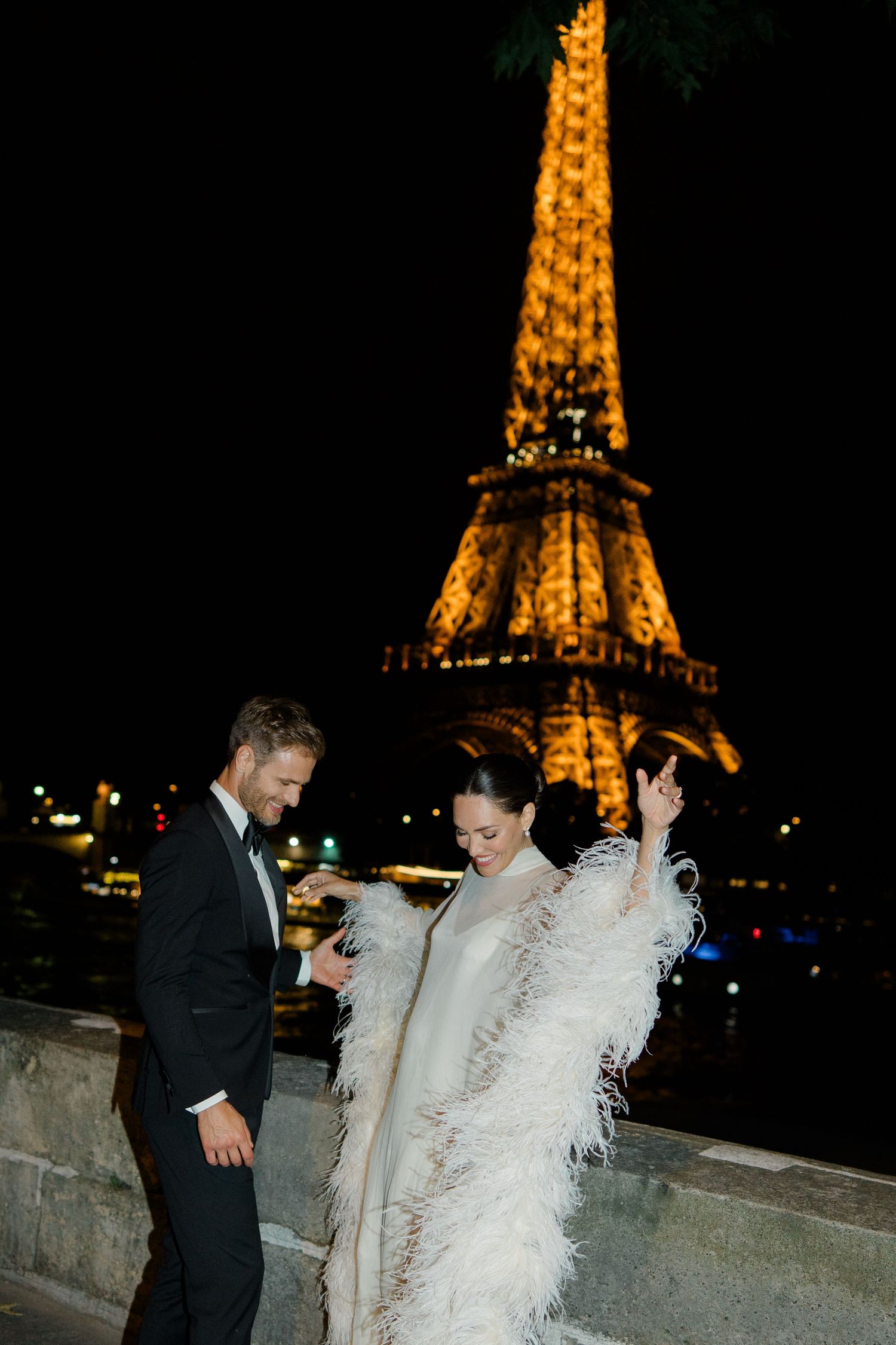 Couple in elegant attire poses before the illuminated Eiffel Tower at night in Paris.