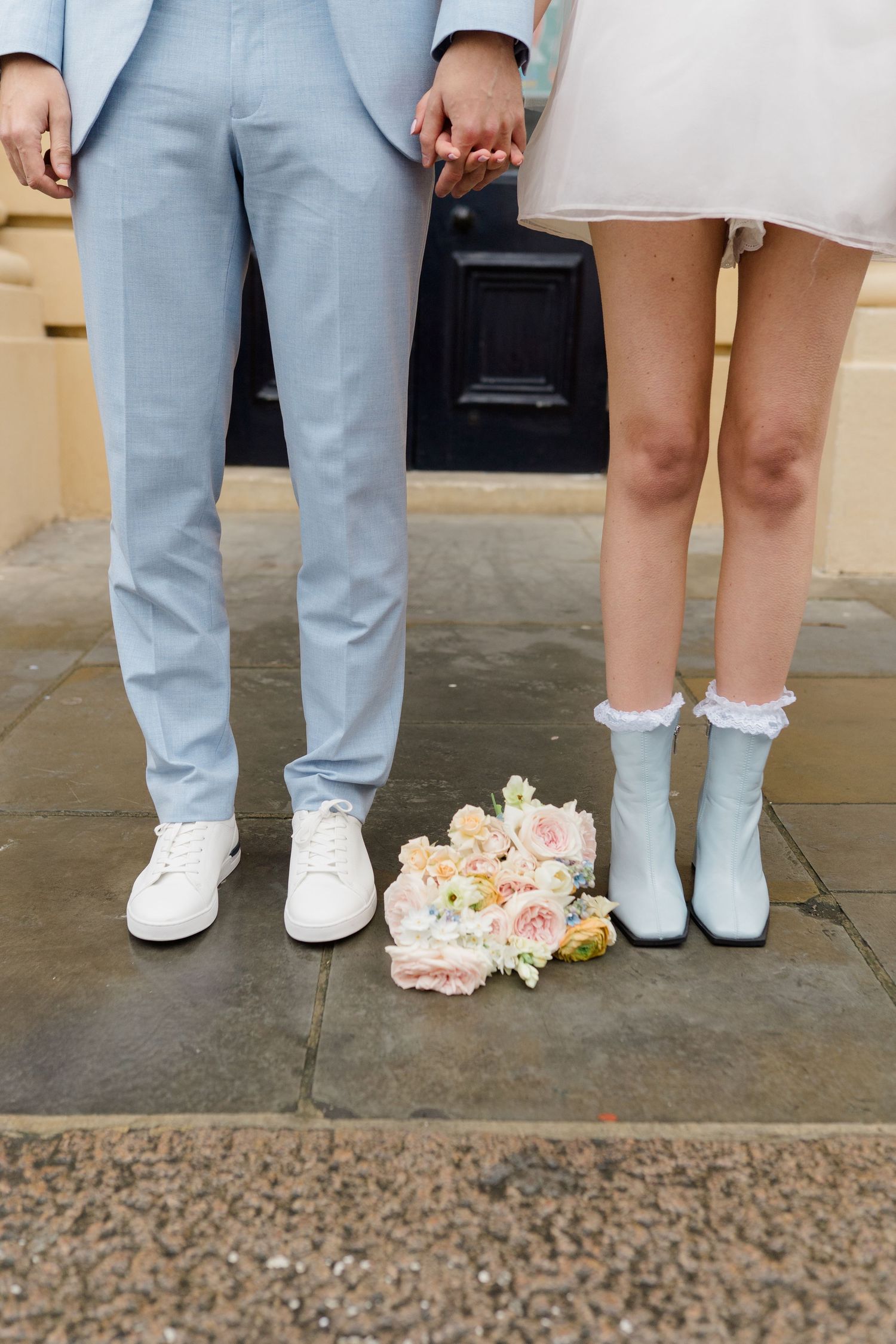 Couple holding hands in wedding attire, blue suit and white mini dress, with bridal bouquet on ground.