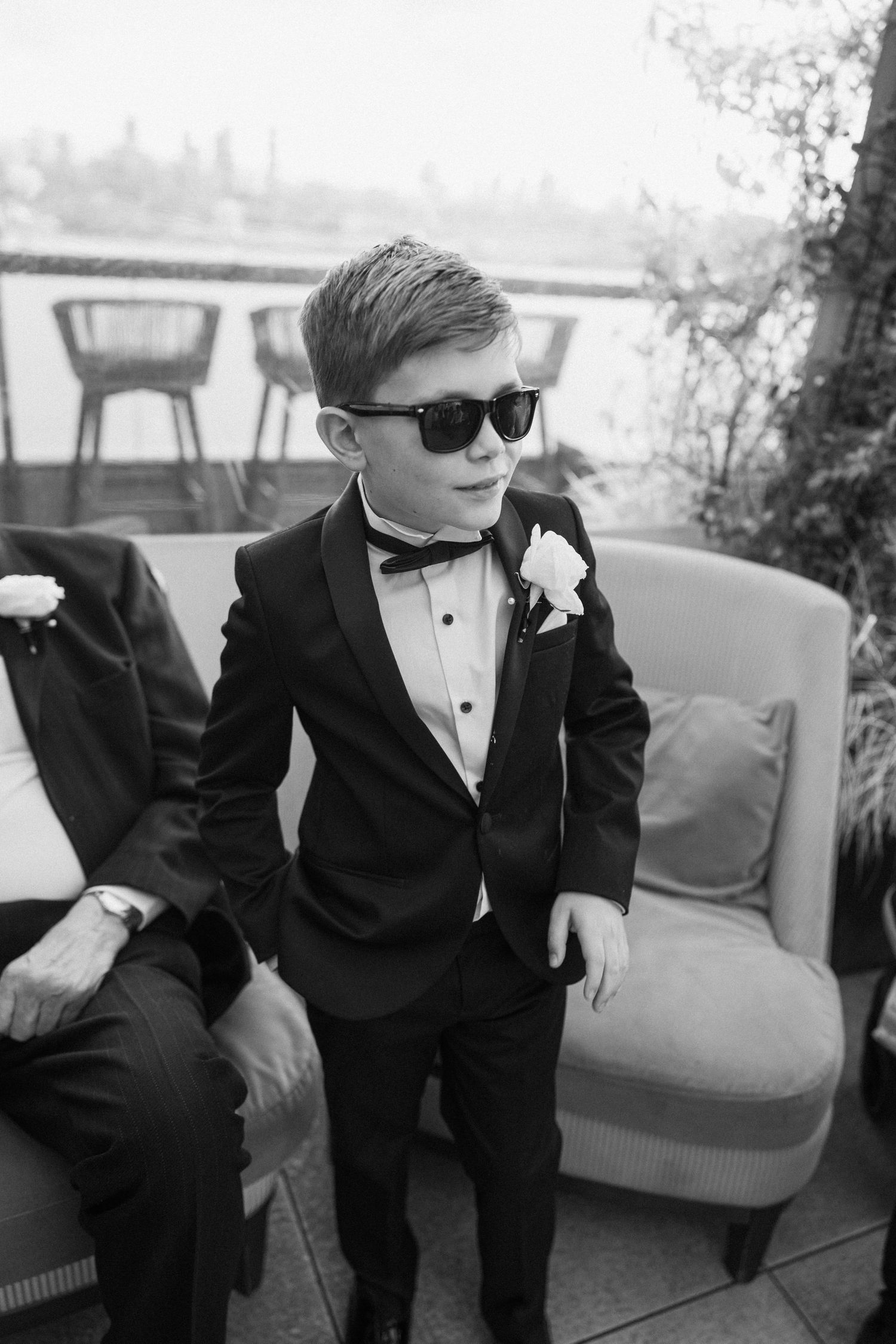 Young ring bearers in formal tuxedos lounging on gray couch at wedding venue.