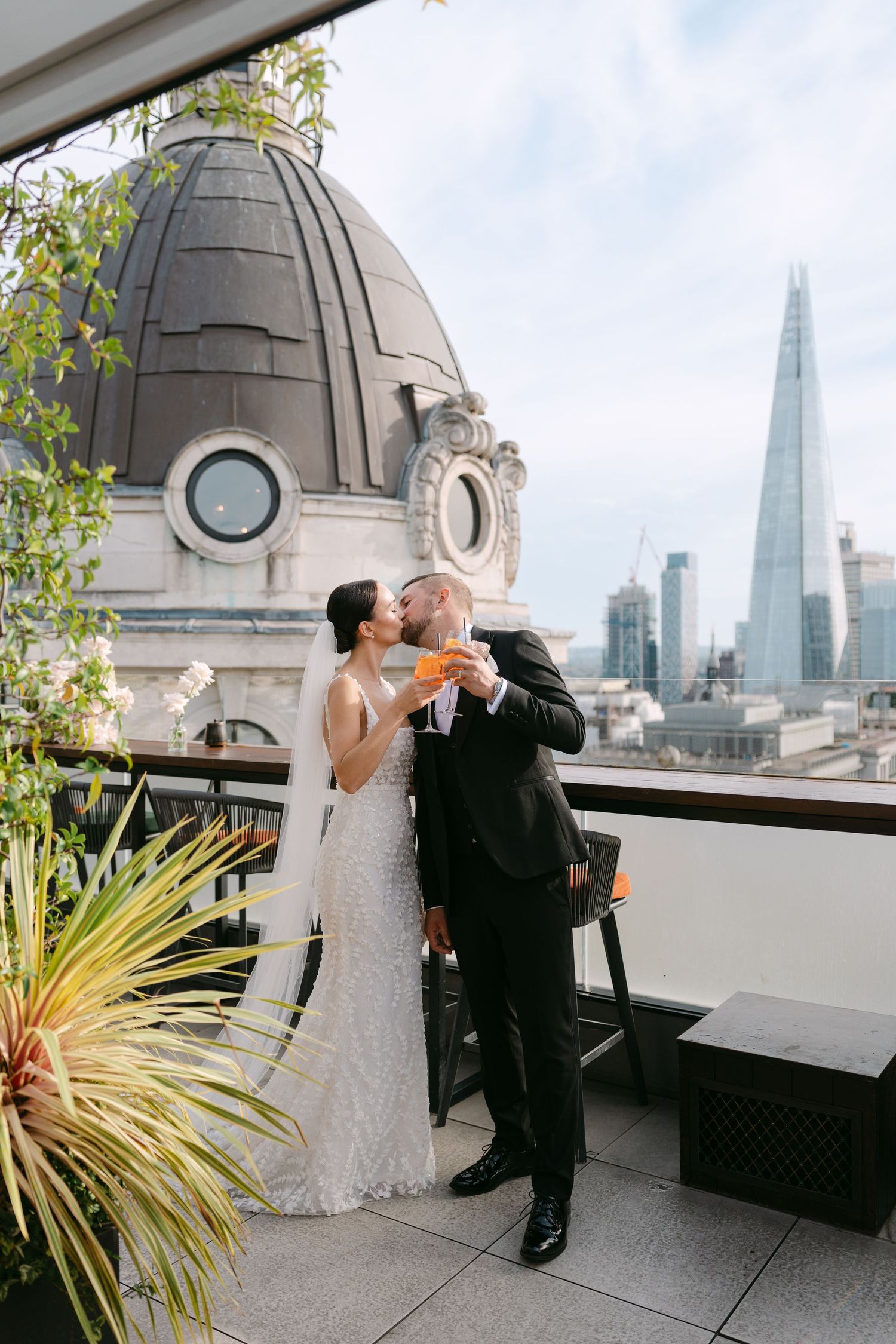 A couple in wedding attire embraces on a rooftop with London's skyline and The Shard visible in the background.