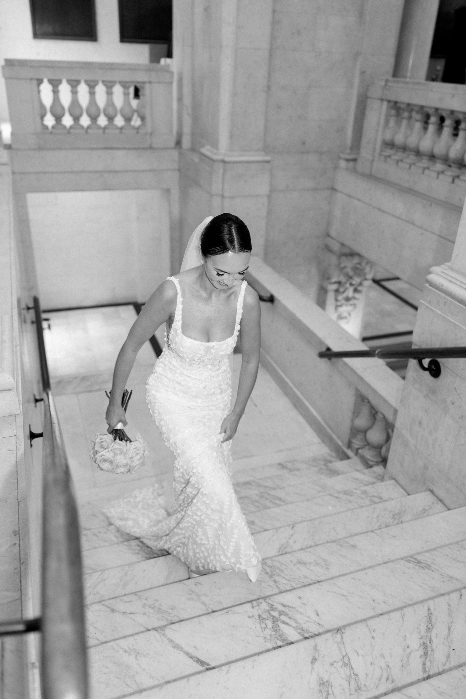 A bride descends marble stairs in an elegant fitted lace wedding gown.