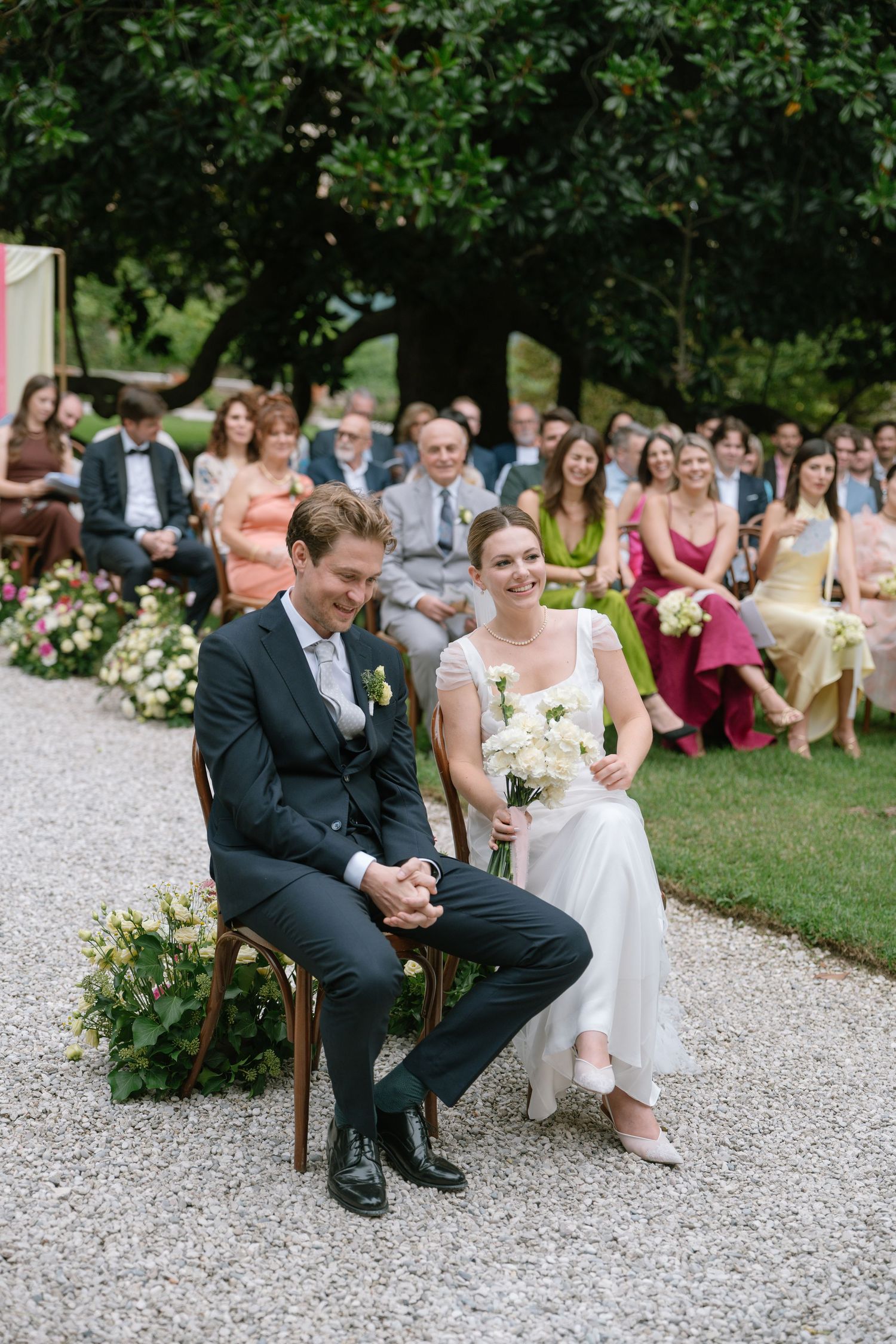 A couple sits on wooden chairs during their outdoor wedding ceremony on a gravel path.