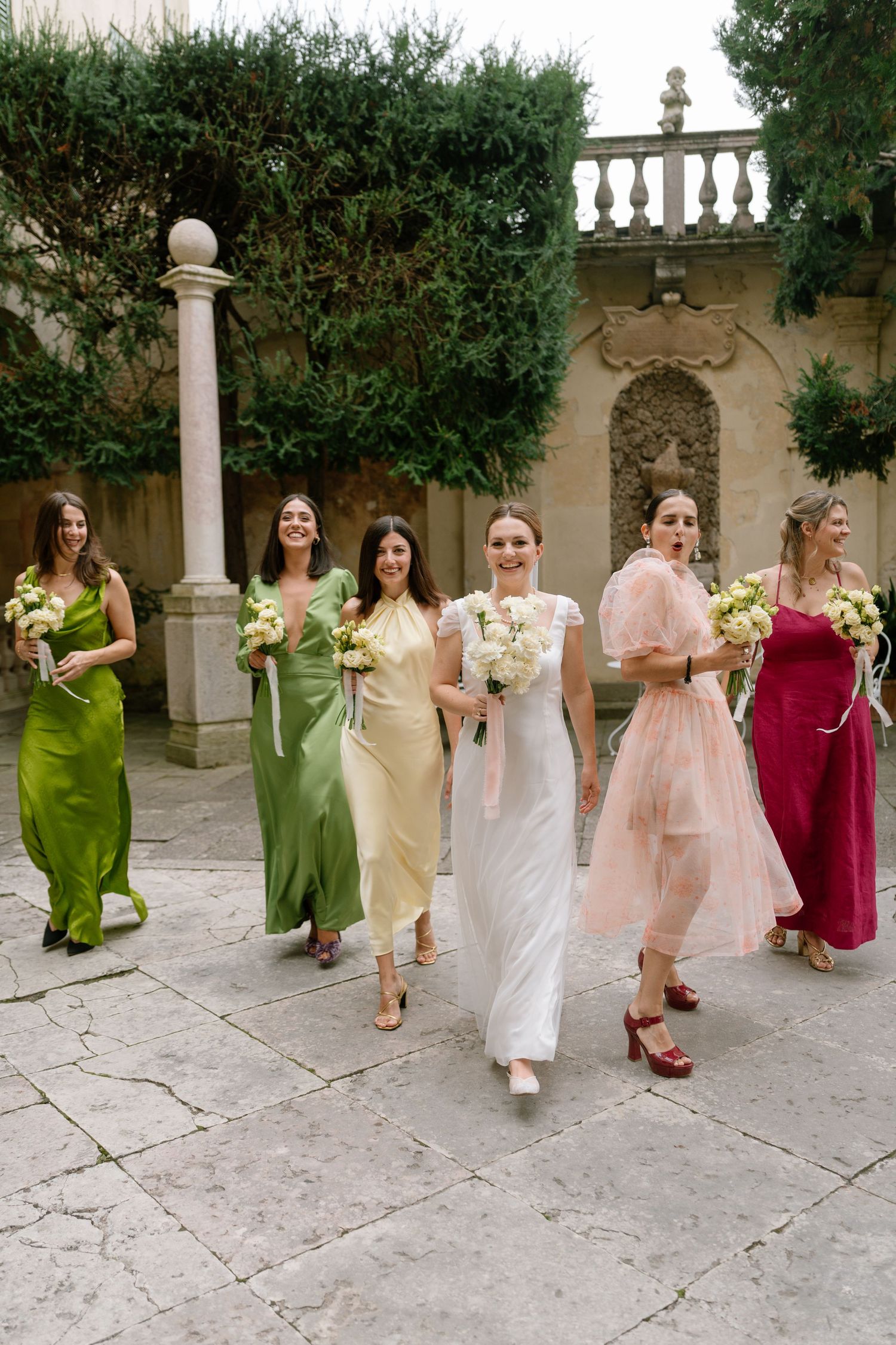 Wedding party in mixed pastel and jewel-toned dresses walks through an ornate garden setting.