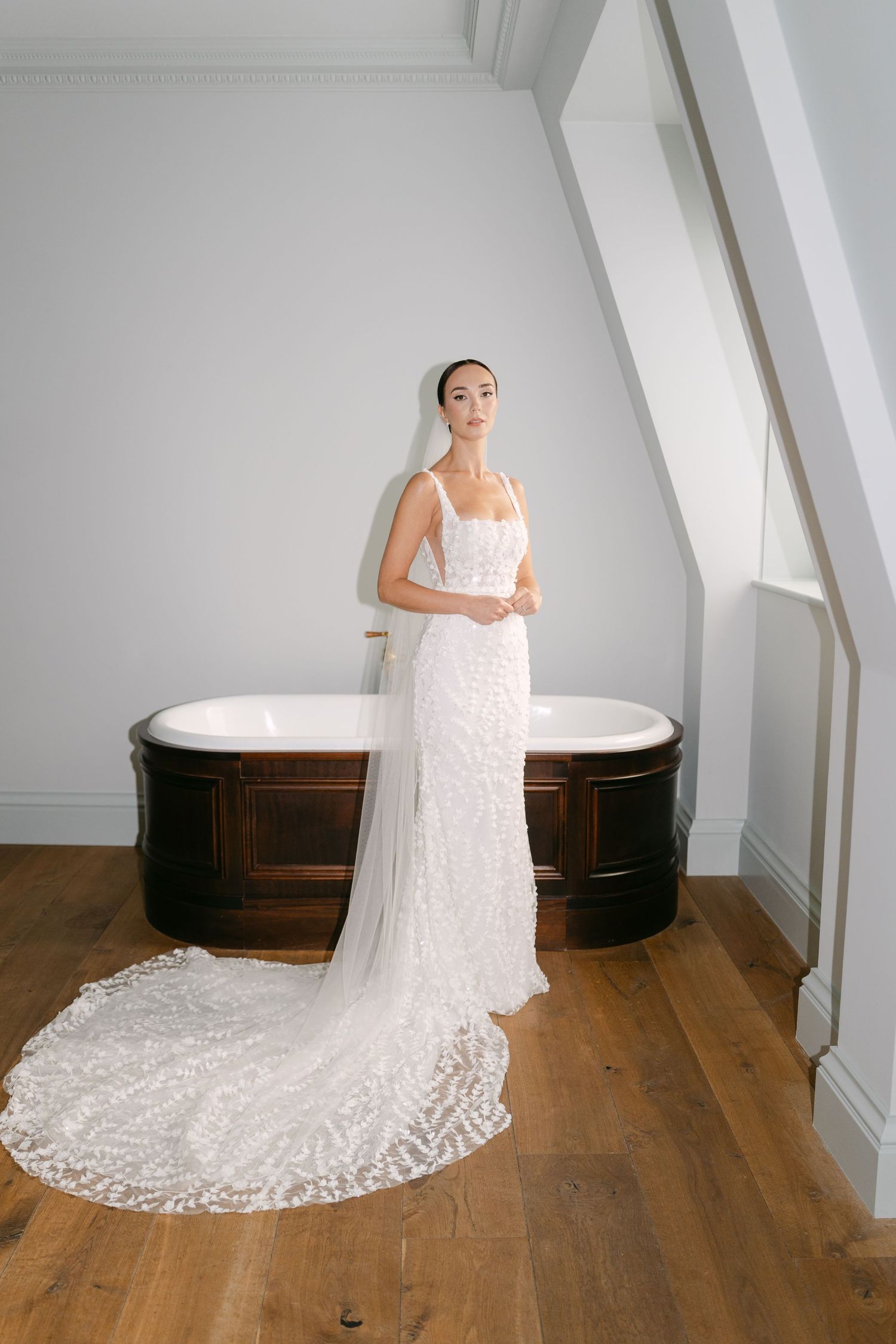 Wedding dress with long lace train displayed in attic room with slanted ceiling and hardwood floors.