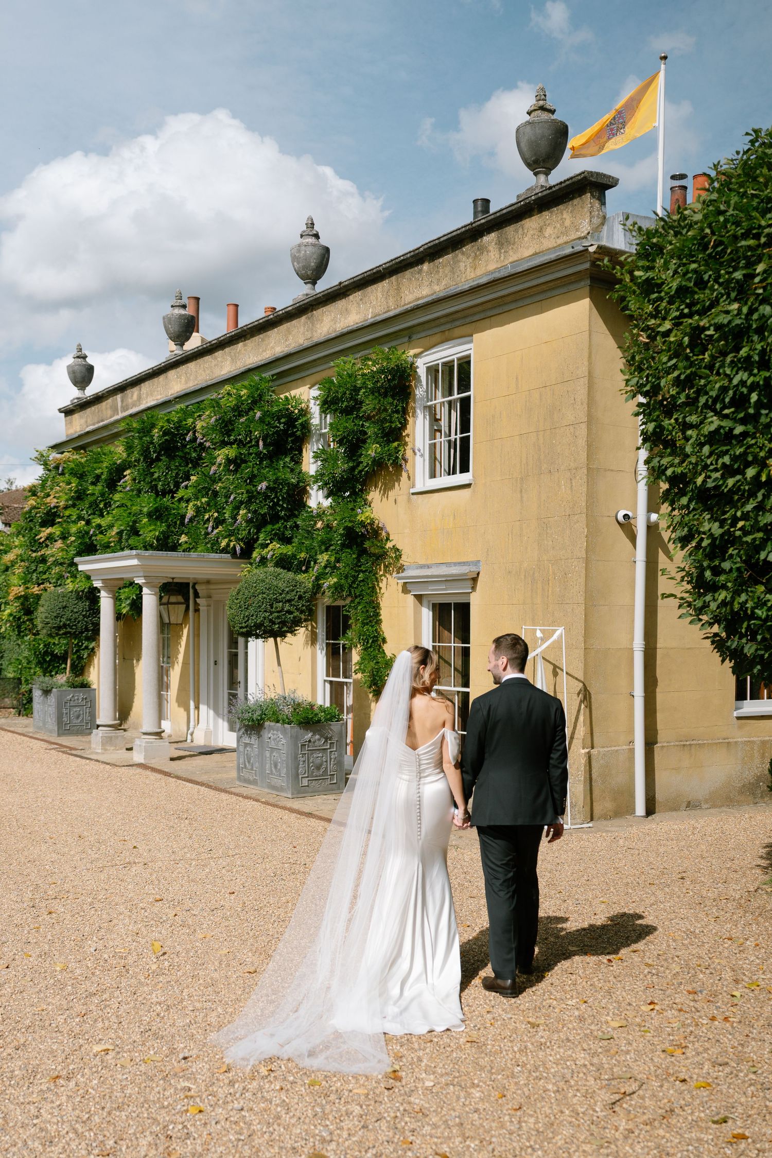 Couple walking along exterior of grand stone building with decorative urns and gravel path.