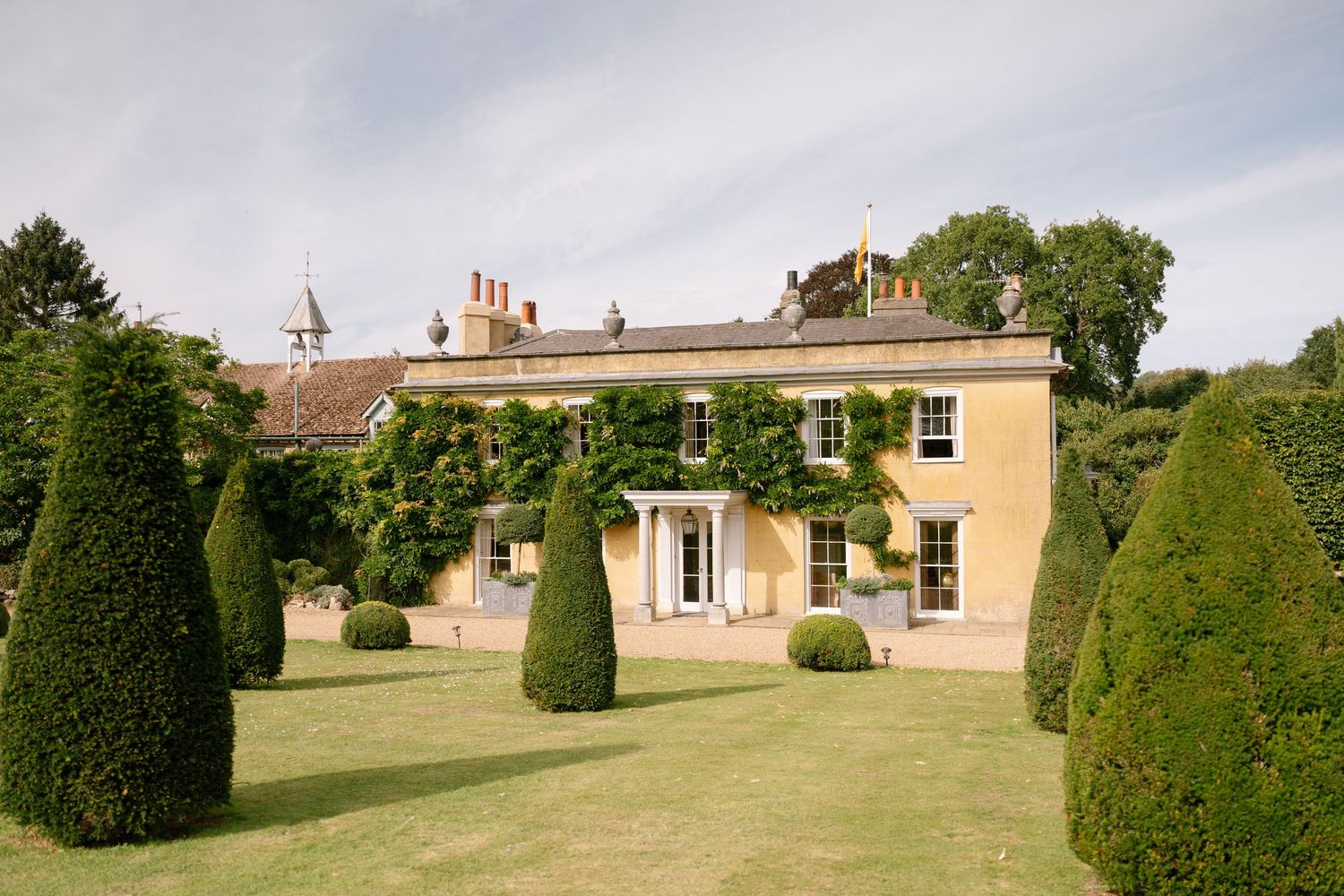 Side view of a yellow manor house with formal topiary gardens and manicured lawn.