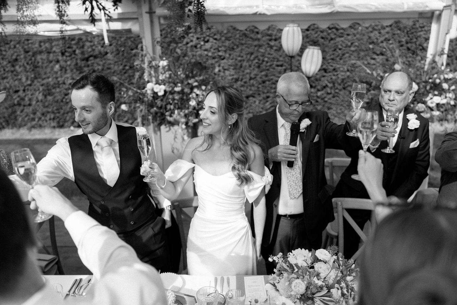 Wedding party raising glasses in a celebratory toast under an outdoor tent.