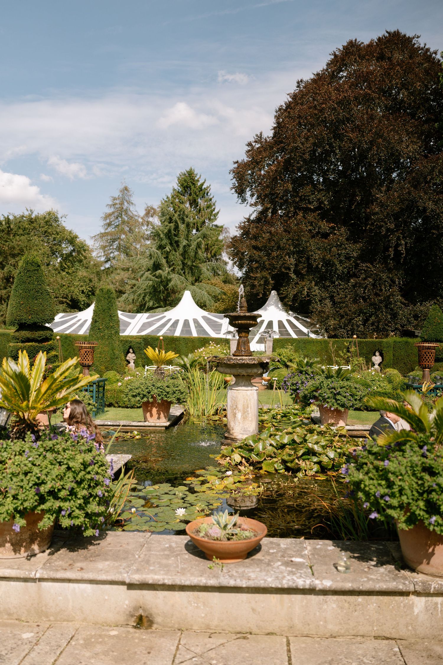 Elegant garden setting with white canopy tents and a water feature surrounded by lush plants.