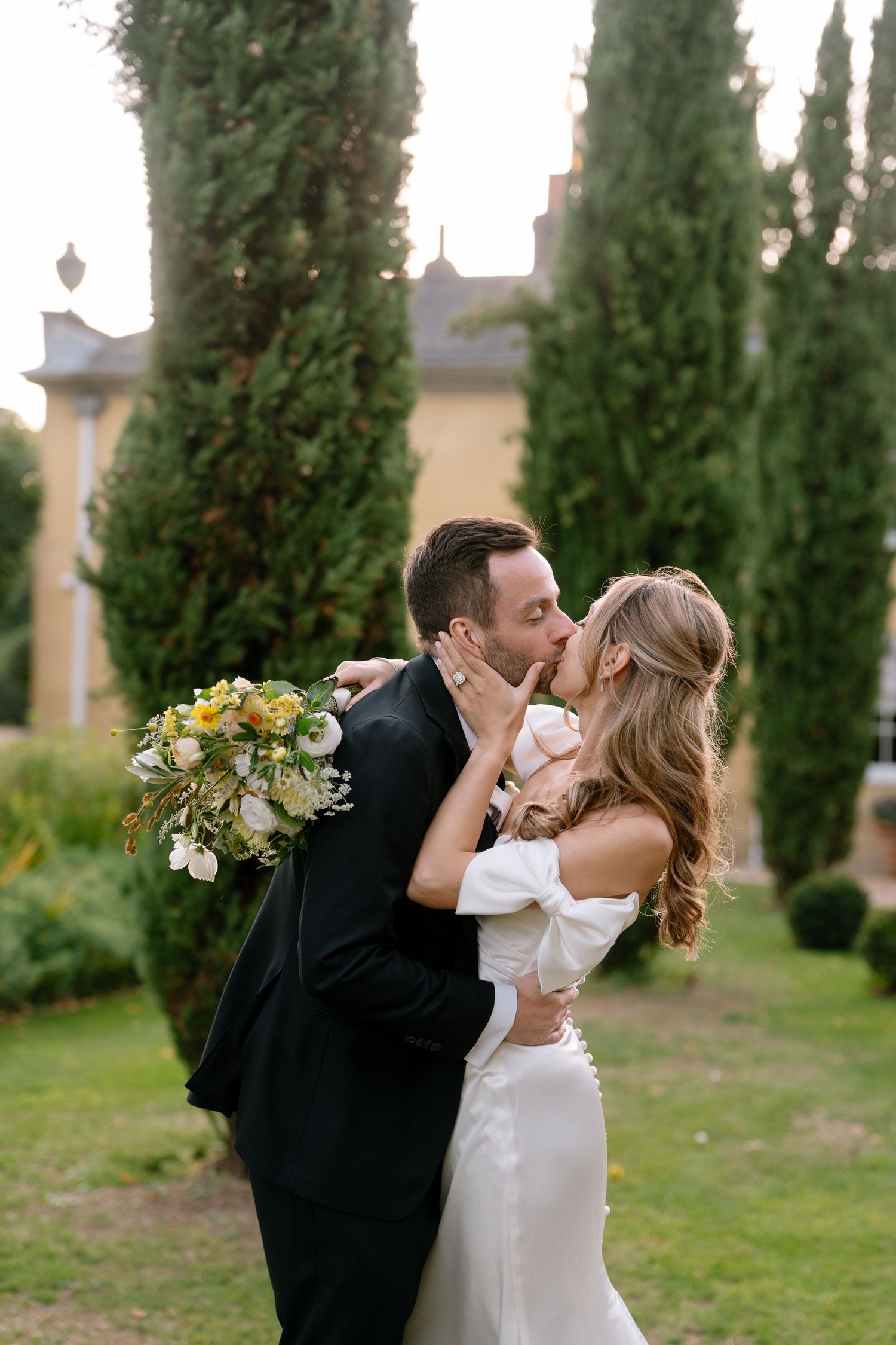 A romantic wedding kiss photographed in a formal garden with cypress trees.