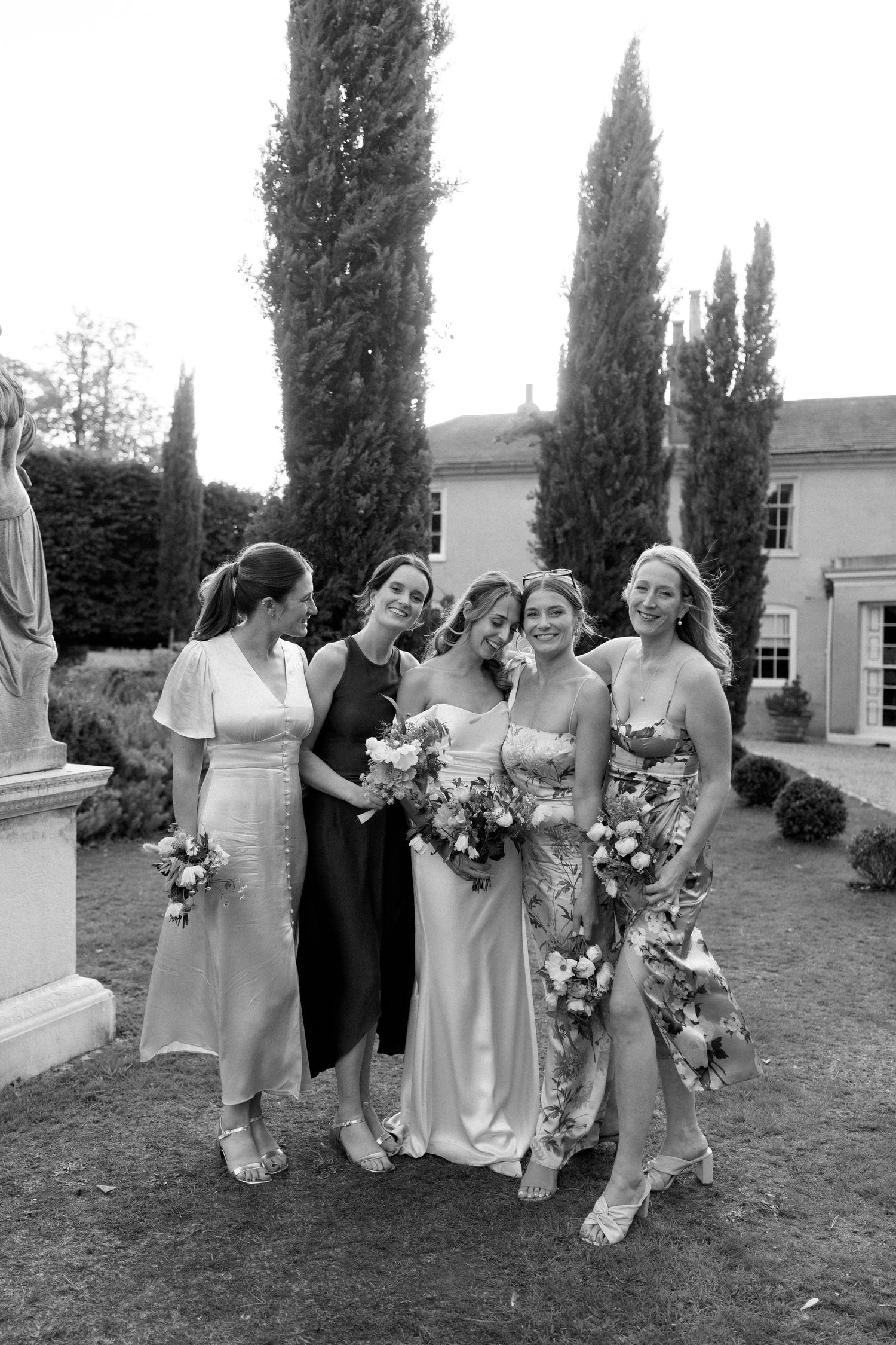 Wedding party poses for candid photos on lawn with white building and cypress trees in background.