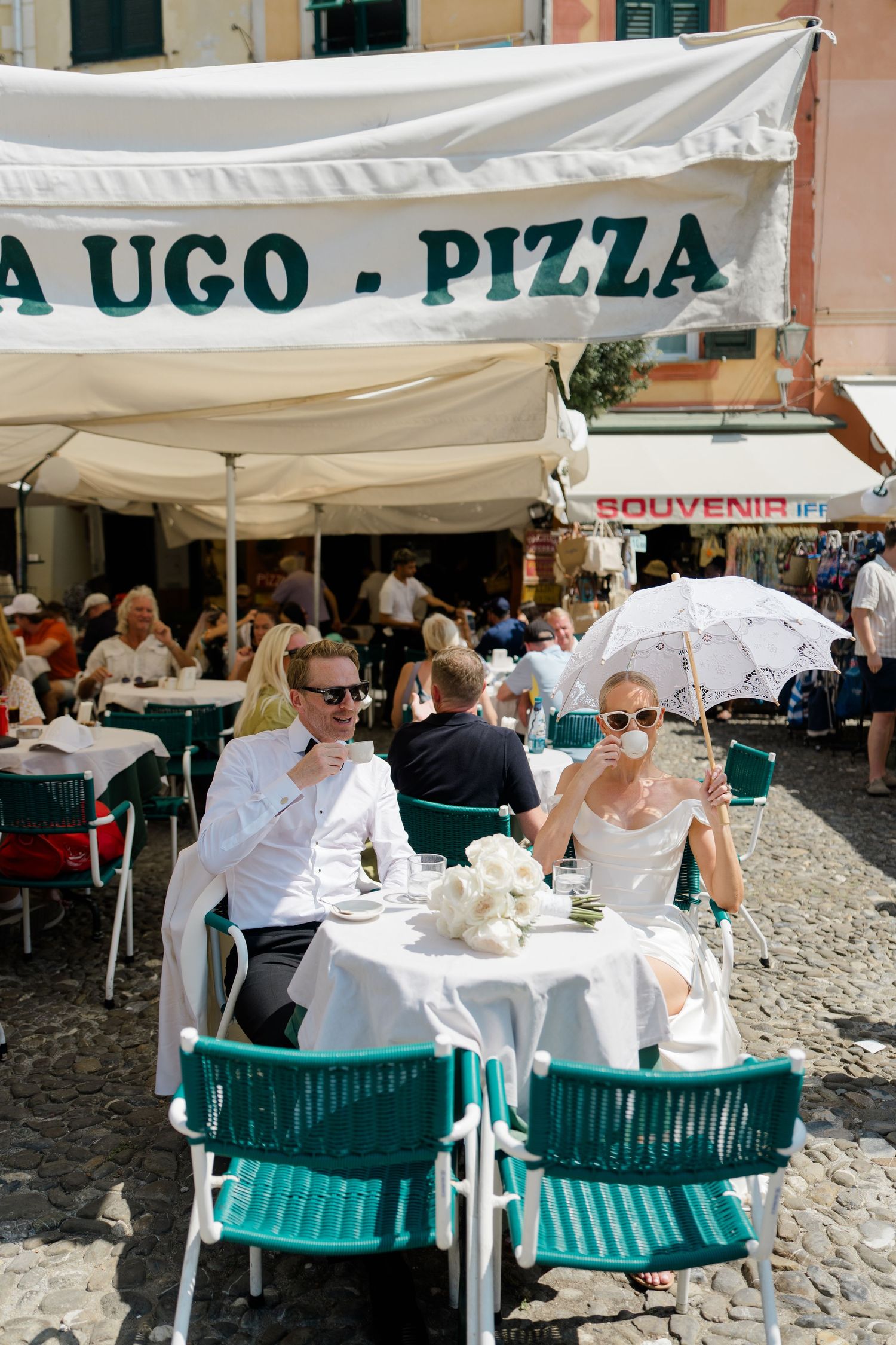 Newlywed couple in white attire celebrating at an outdoor pizza restaurant in a sunny Italian piazza.