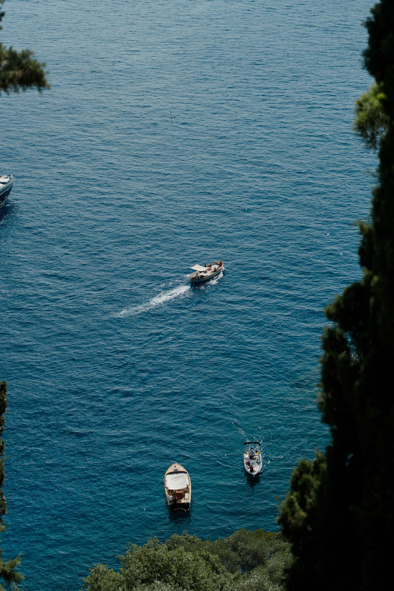 Aerial view of small boats floating on vivid blue Mediterranean waters framed by lush green trees.