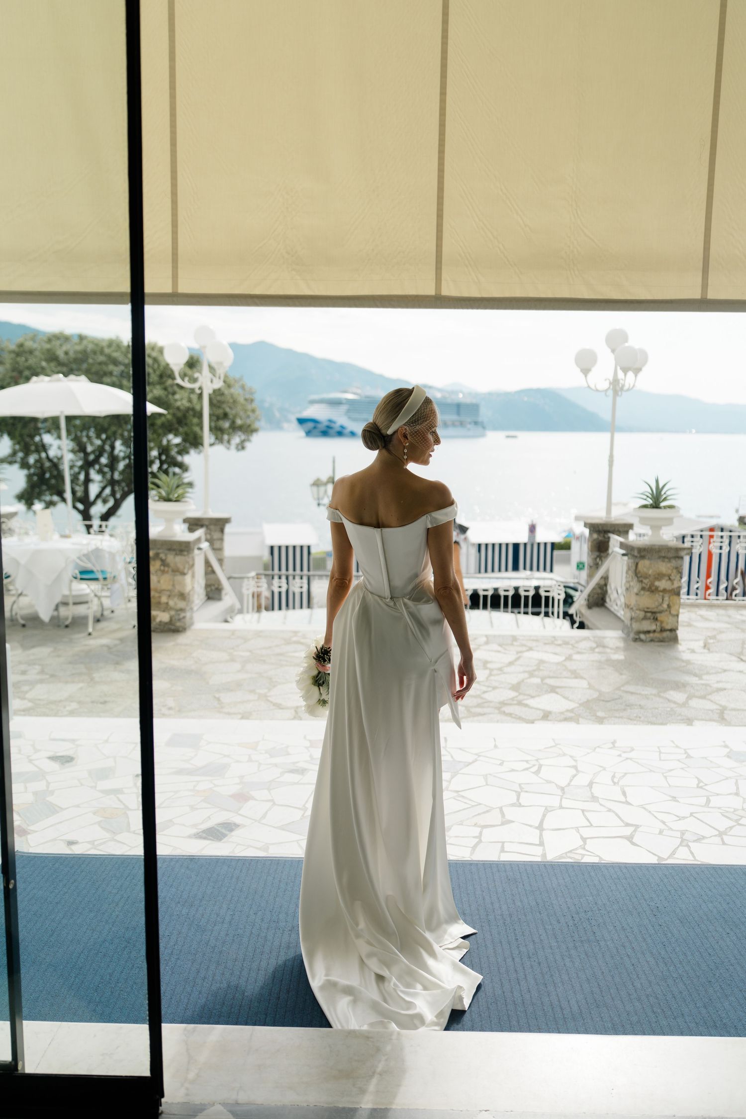 Bride in off-shoulder white gown with headpiece stands at glass doors overlooking a seaside terrace.