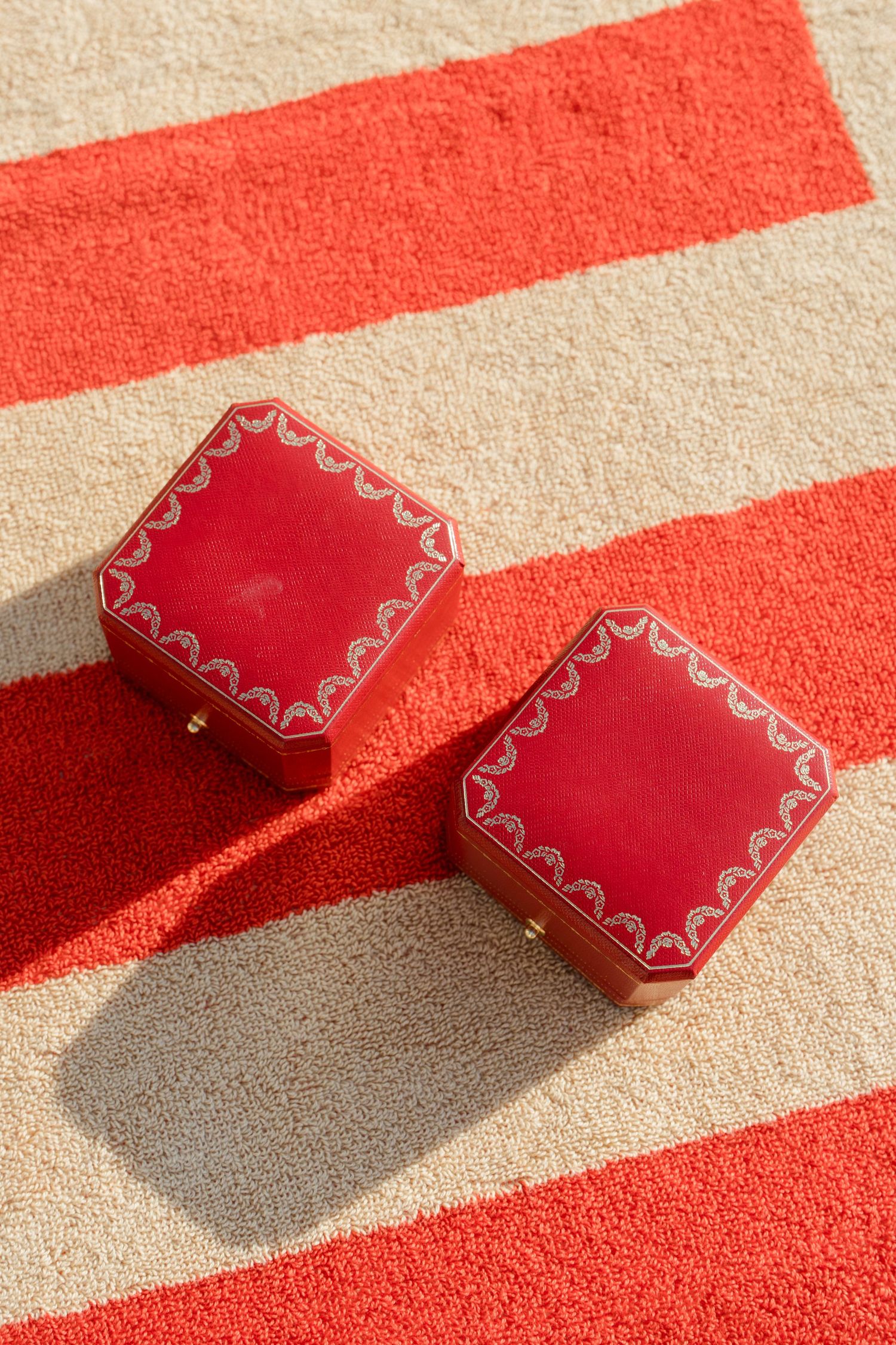 Two red Cartier jewelry boxes with ornate gold detailing resting on a striped red and cream towel in sunlight.